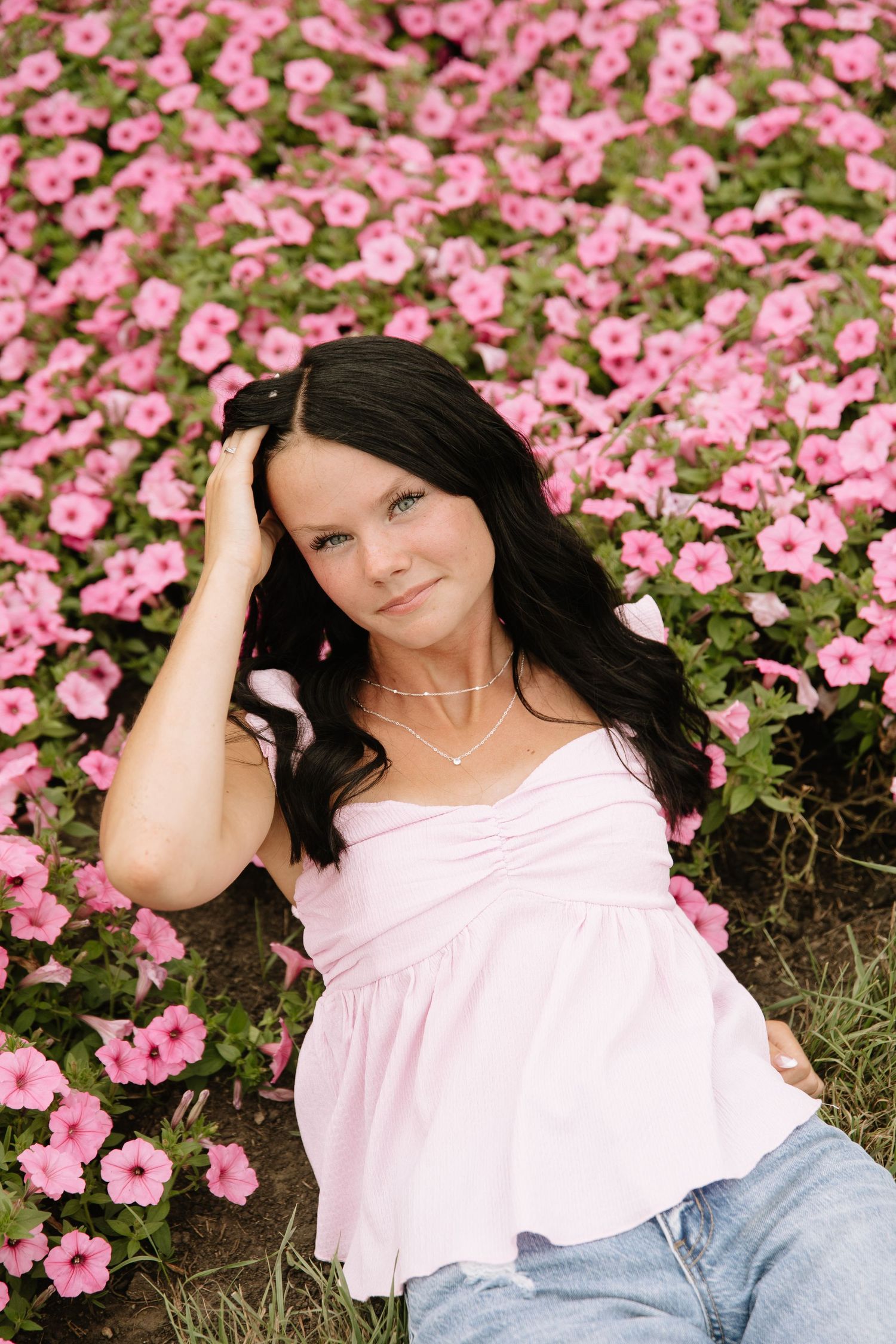 Young woman with dark hair in pink top sits among blooming pink petunias, smiling at camera.