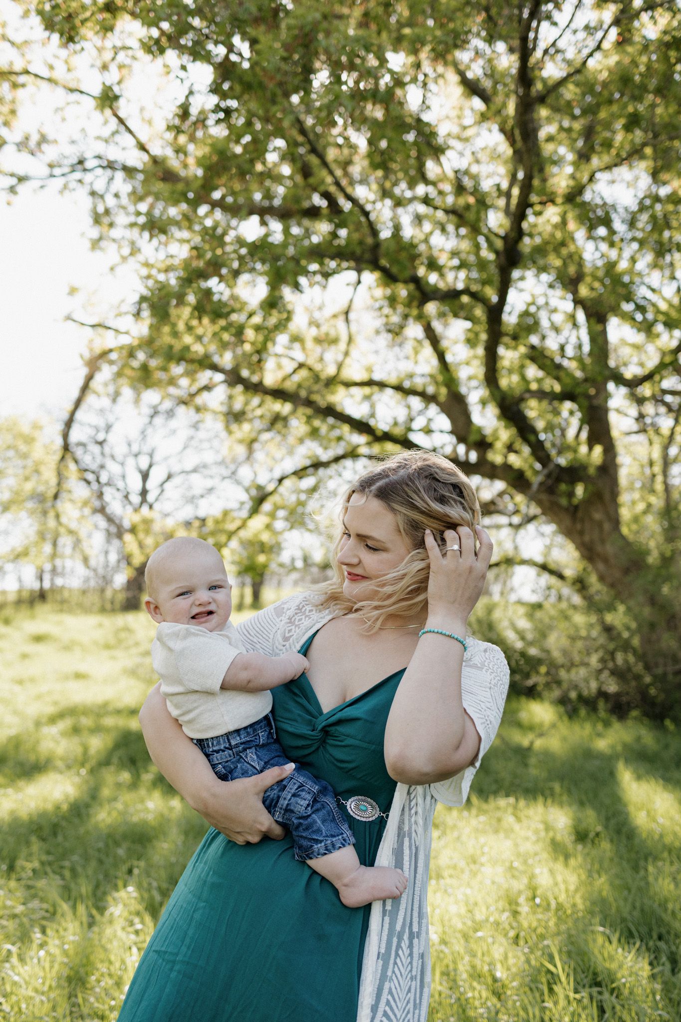 A young blonde woman in a teal dress holds her baby boy outdoors in a lush green field beneath a large tree.
