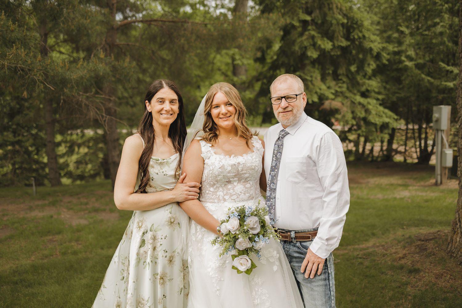 Bride in white gown holding bouquet poses with two guests in a lush green outdoor setting.