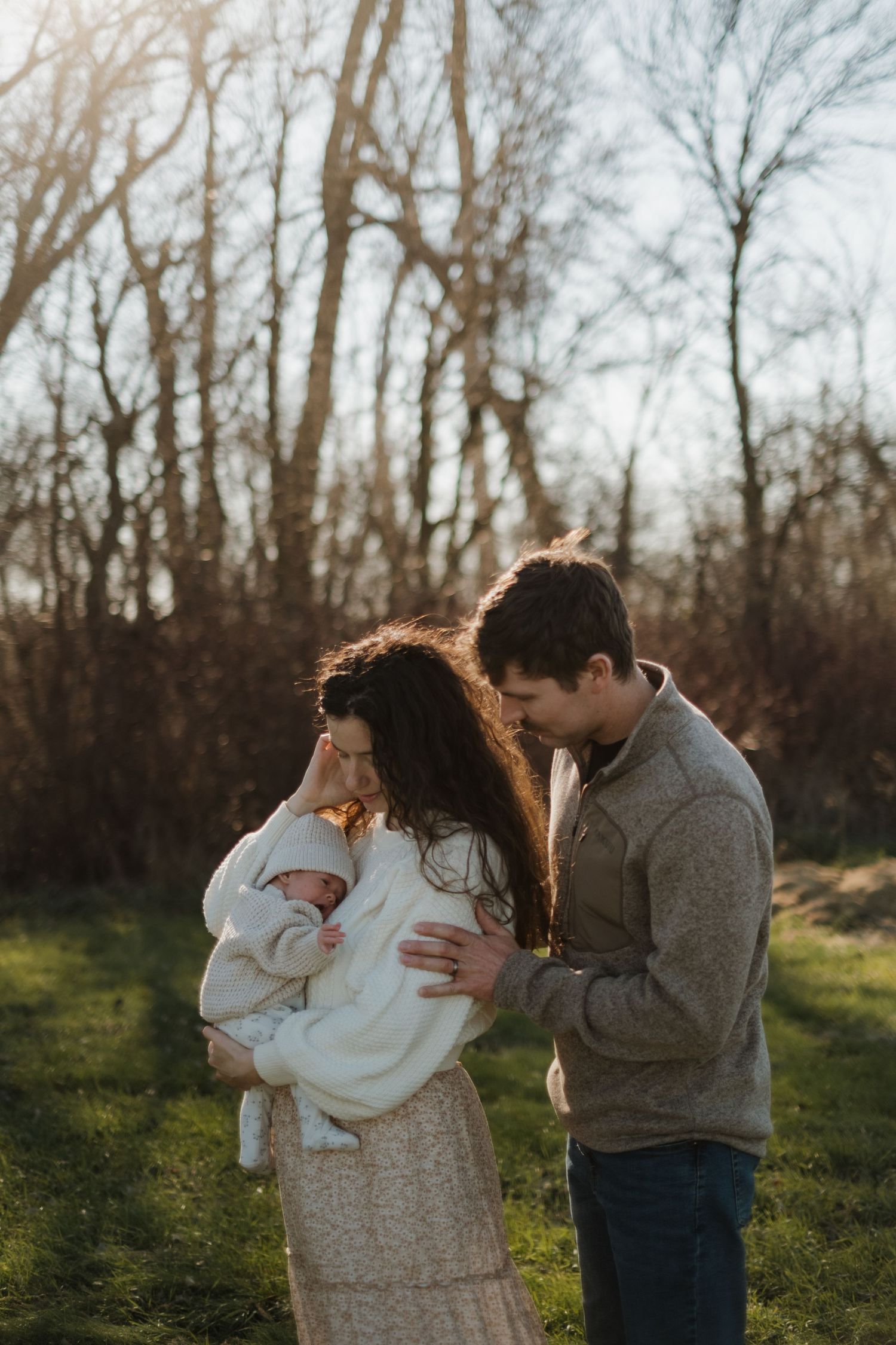 Family in white and gray clothing embracing their baby during golden hour in a wooded setting.