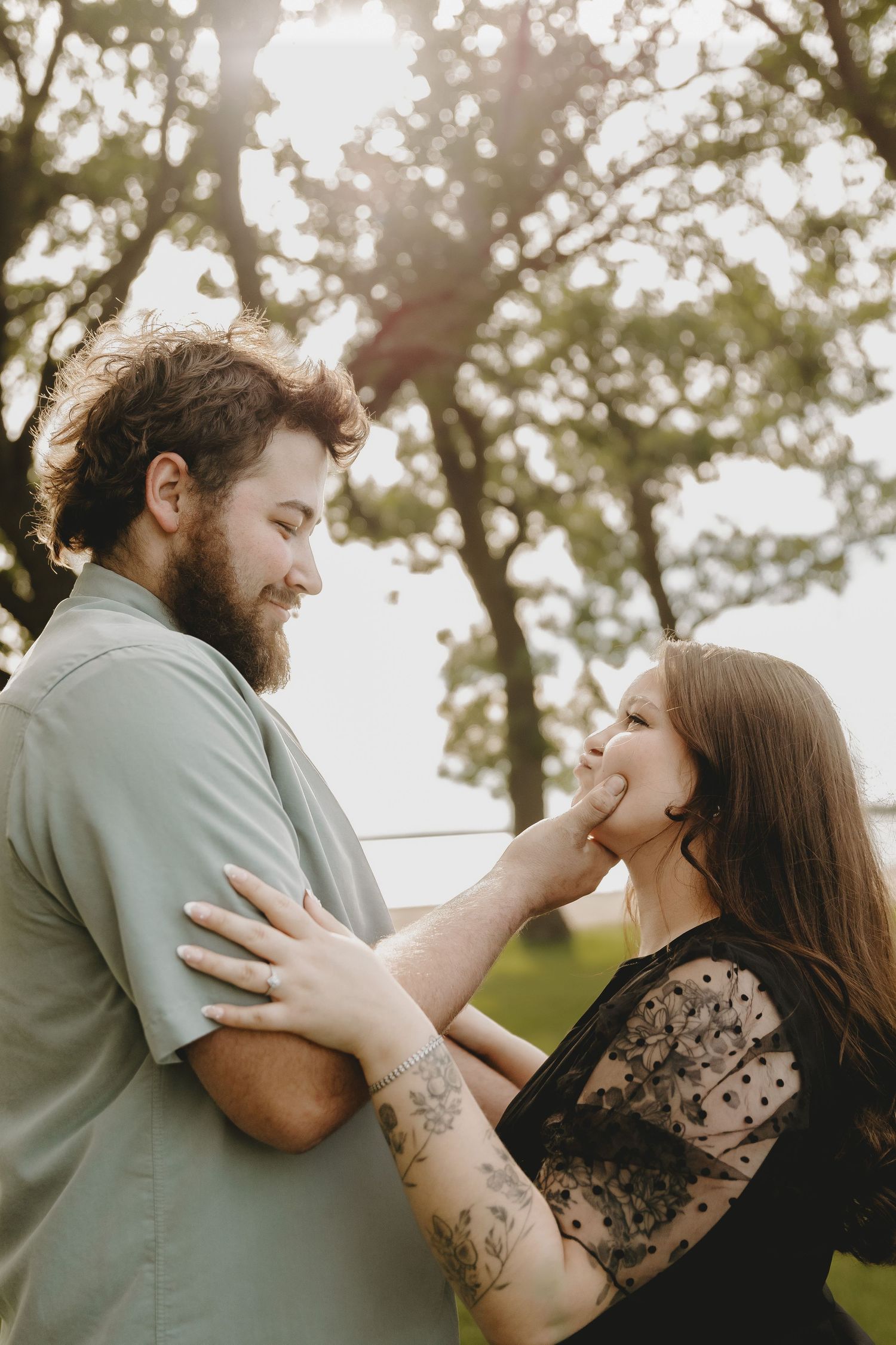 A bearded man tenderly cups a smiling woman's face outdoors, with warm sunlight filtering through green trees.