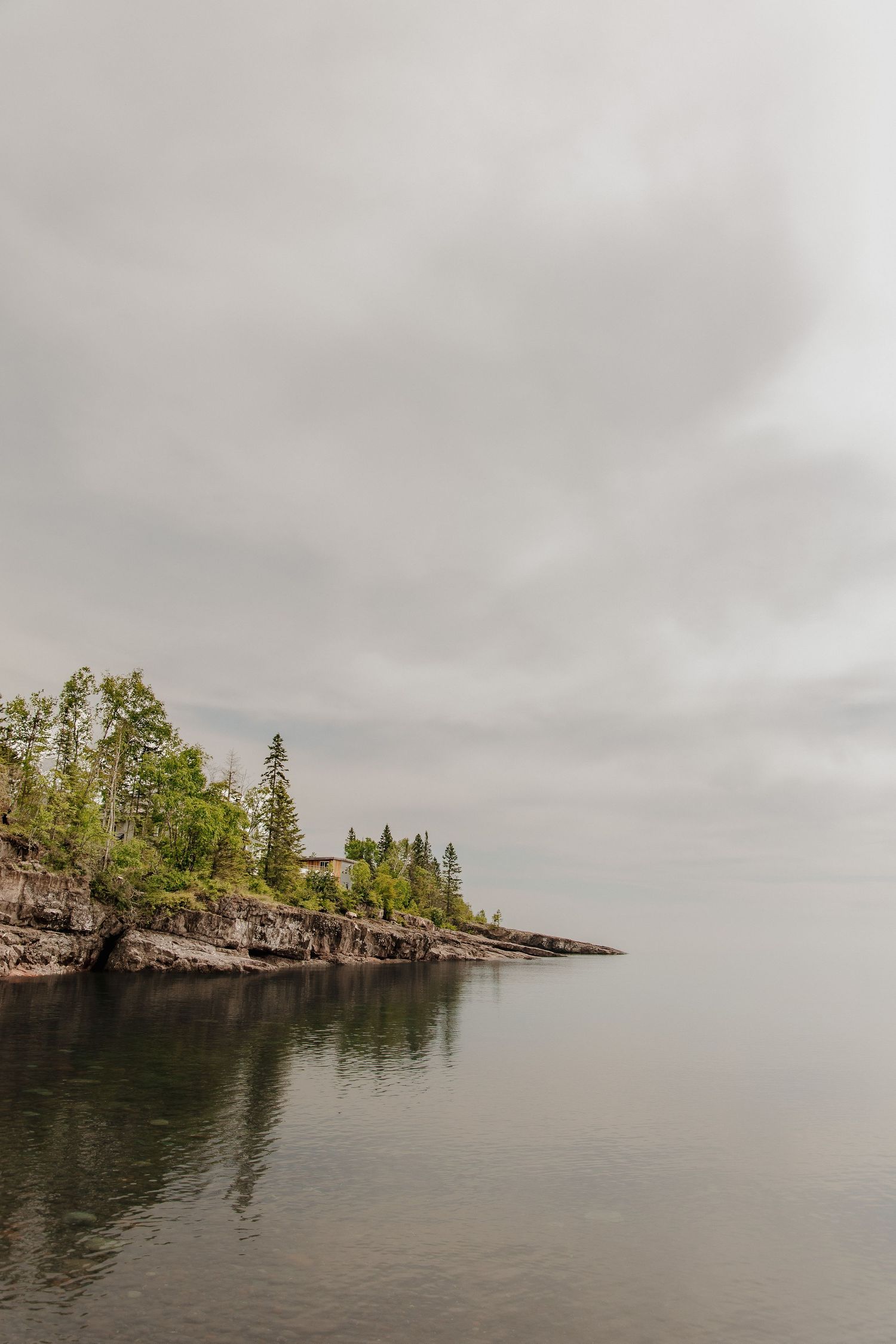 Rocky shoreline with evergreen trees under overcast skies reflecting on calm lake waters.