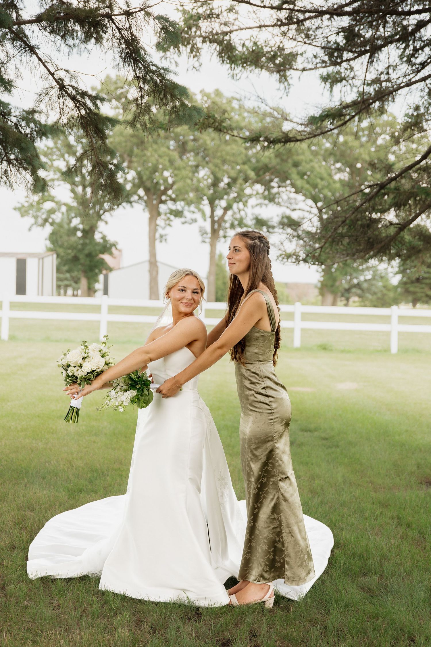 Bride in strapless gown holding bouquet dancing with bridesmaid in olive dress under pine tree.