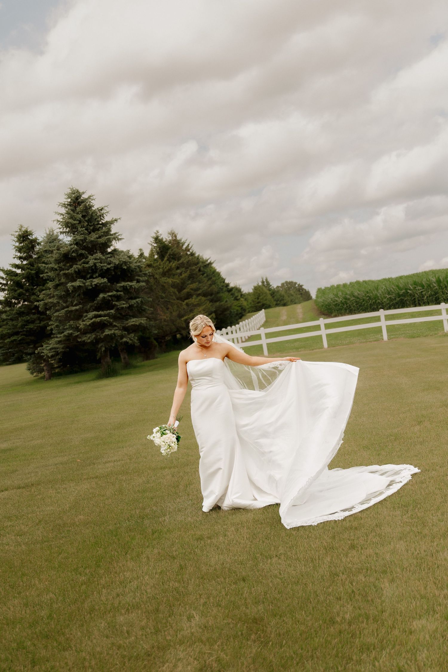 Bride in flowing white gown twirling on green lawn with white fence and evergreen trees backdrop.