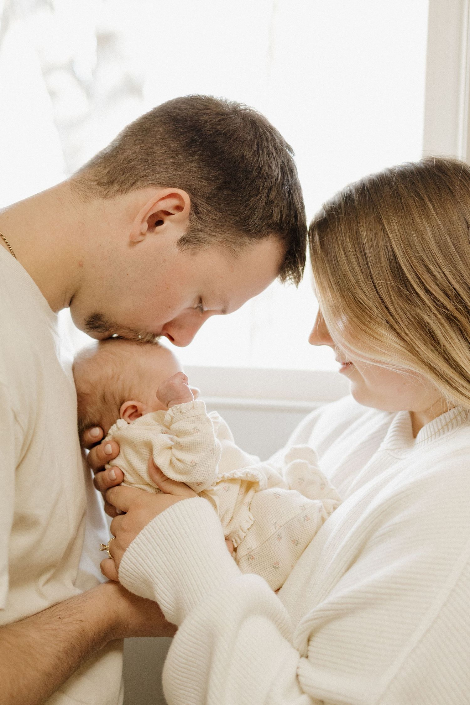 Parents tenderly kissing their newborn baby, dressed in matching cream outfits, in a bright and airy room.