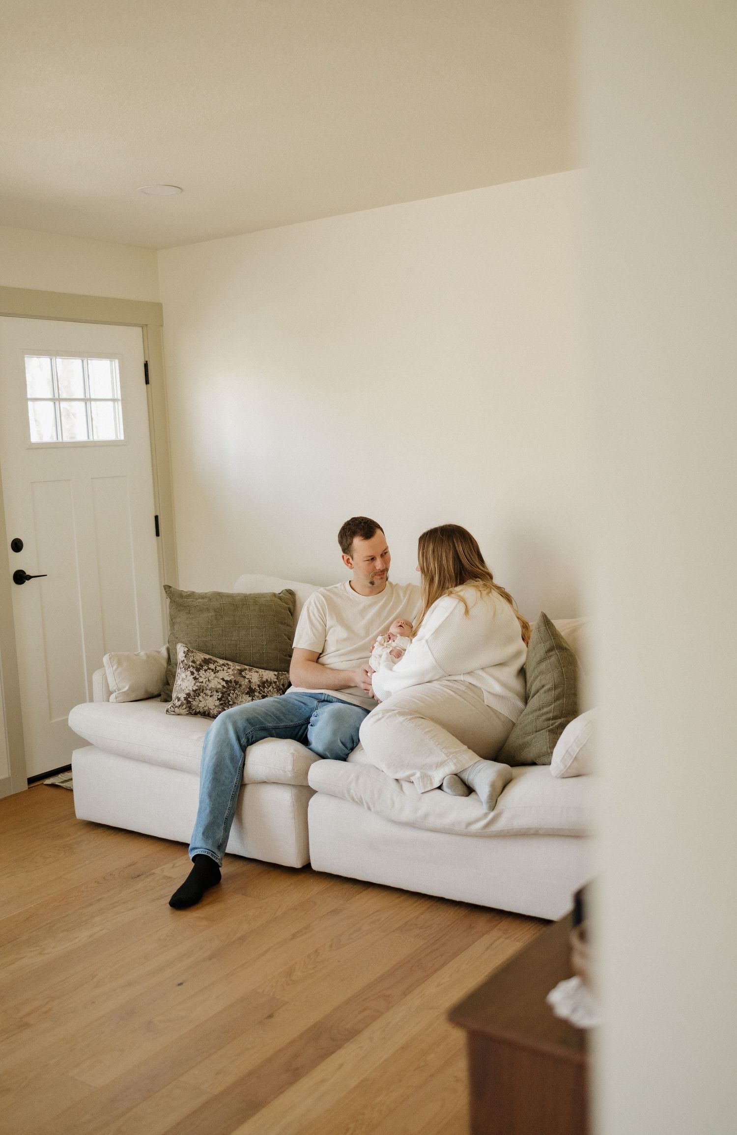 A couple sits on a white sofa in a bright living room, sharing a tender moment with their newborn baby.
