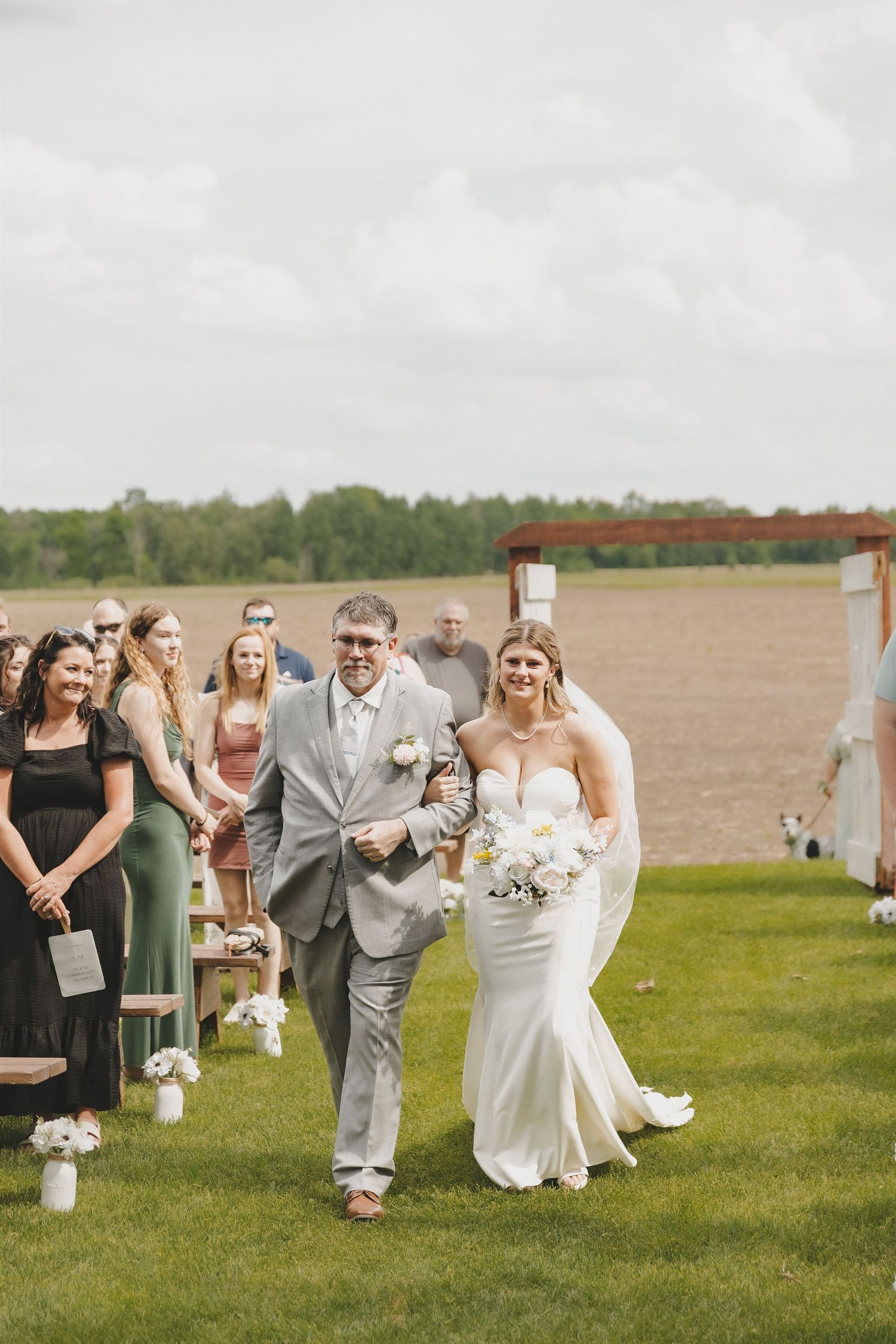 Father walks smiling bride in white mermaid gown down outdoor aisle at rustic wedding.