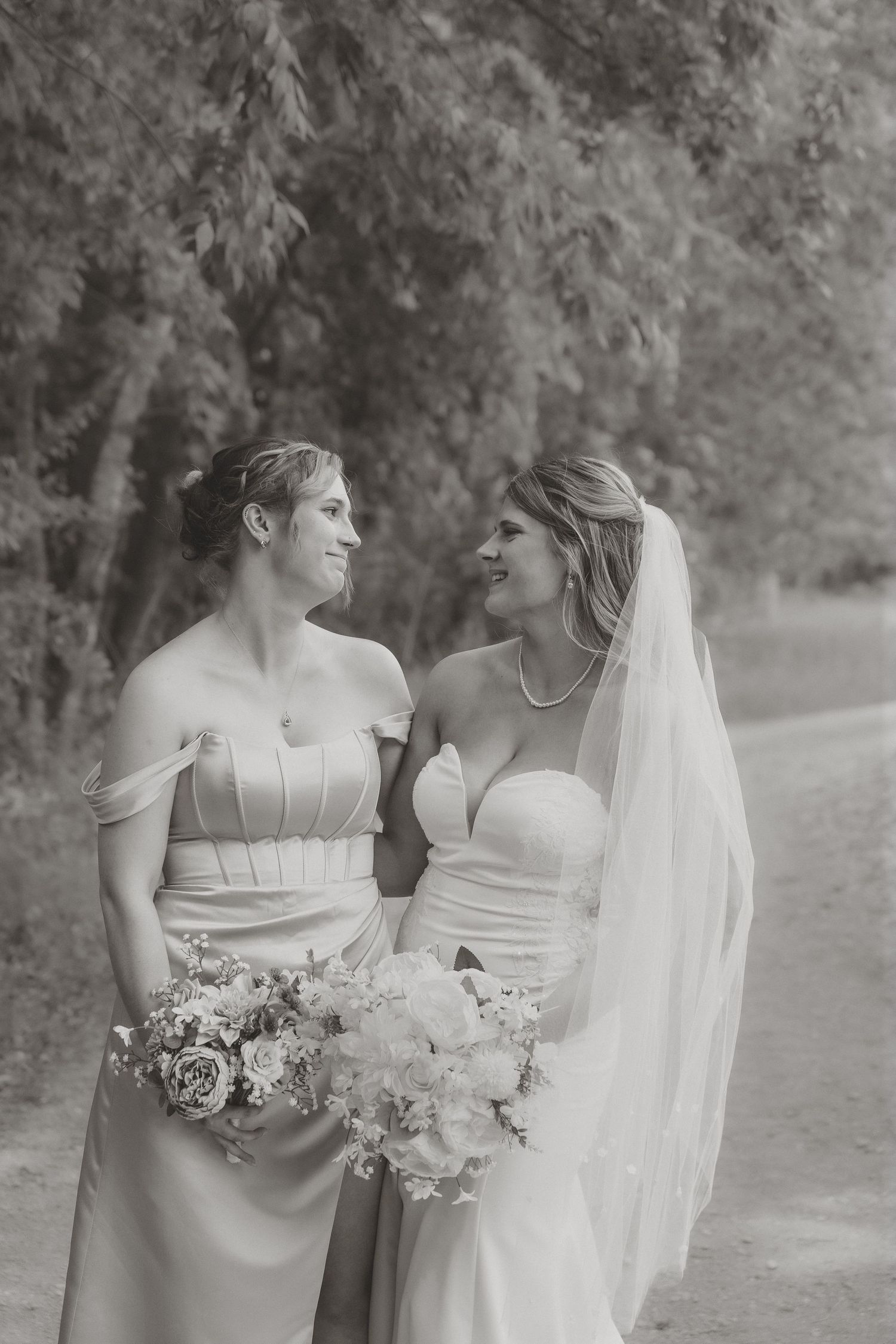 Black and white photo of bride and bridesmaid smiling at each other holding bouquets outdoors.
