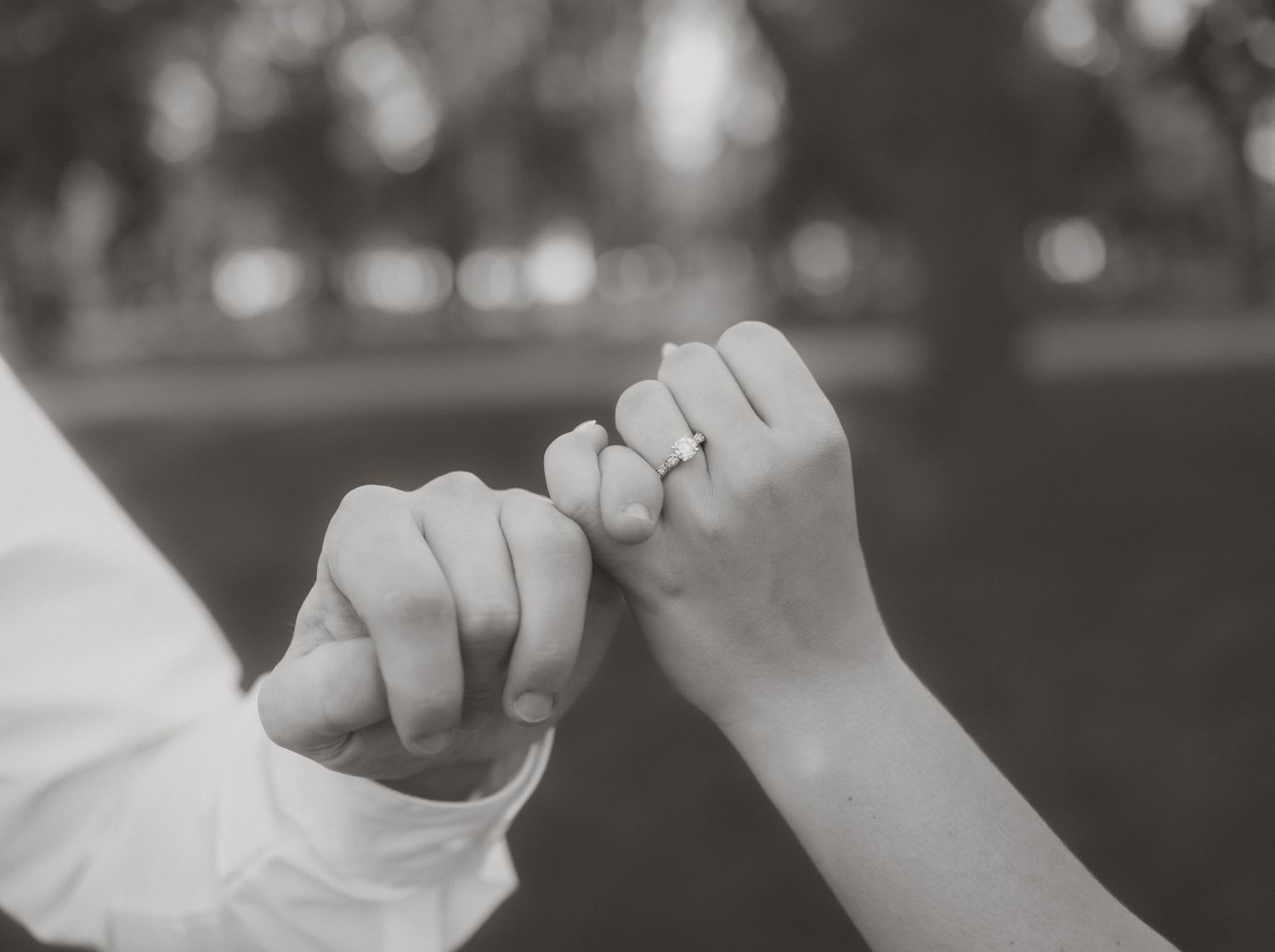 Black and white photo of a couple doing a pinky promise, woman wearing an engagement ring outdoors.