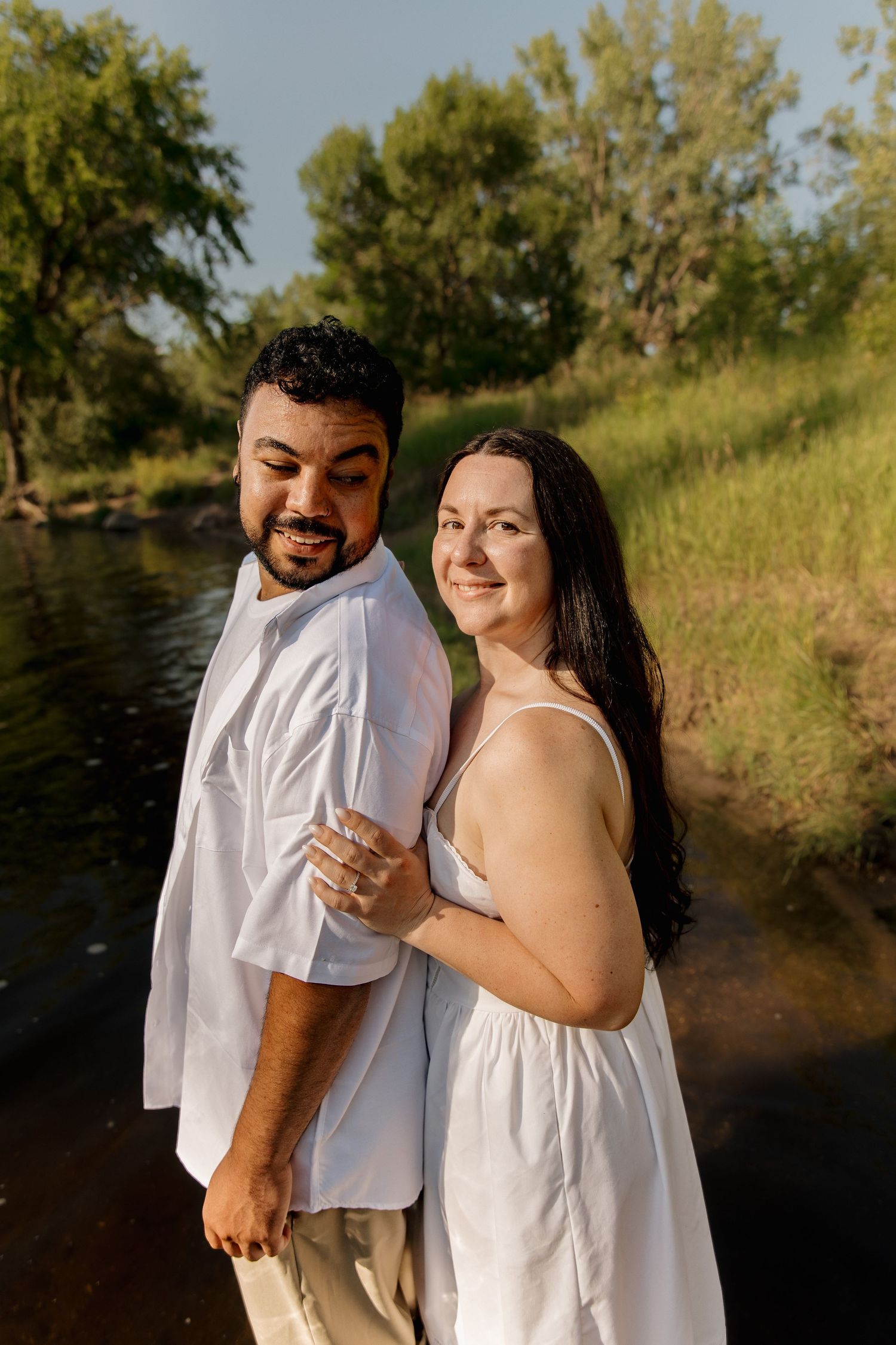 A smiling couple in white outfits poses by a sunlit river surrounded by lush green trees and grass.