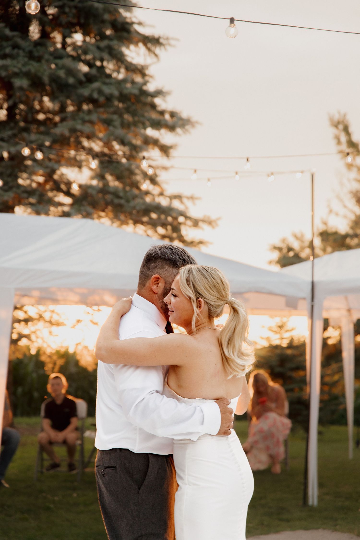 Bride and groom share romantic first dance embrace at golden hour outdoor wedding reception under lights.