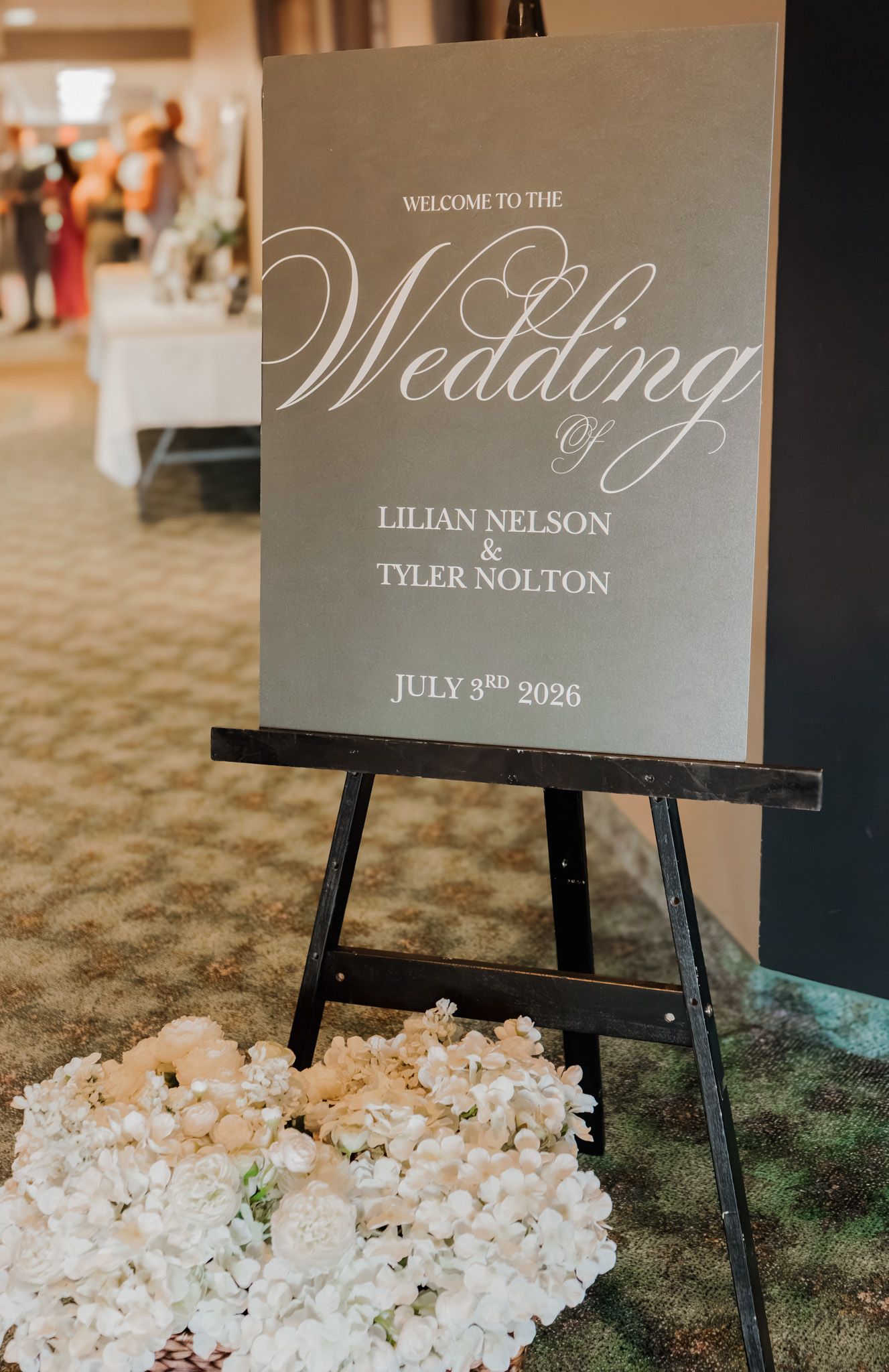 Elegant wedding welcome sign on easel for Lilian Nelson and Tyler Nolton, July 3rd 2026, with white floral arrangement.