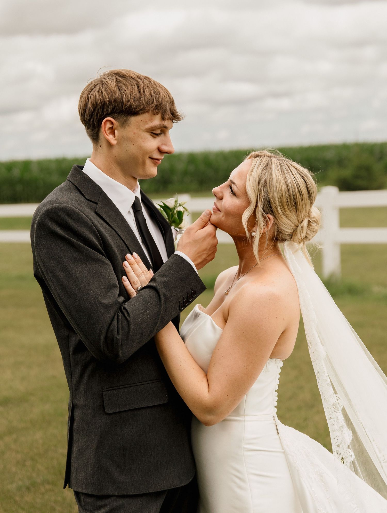 A groom in a dark suit tenderly touches his brides chin as she smiles in a white strapless gown outdoors.