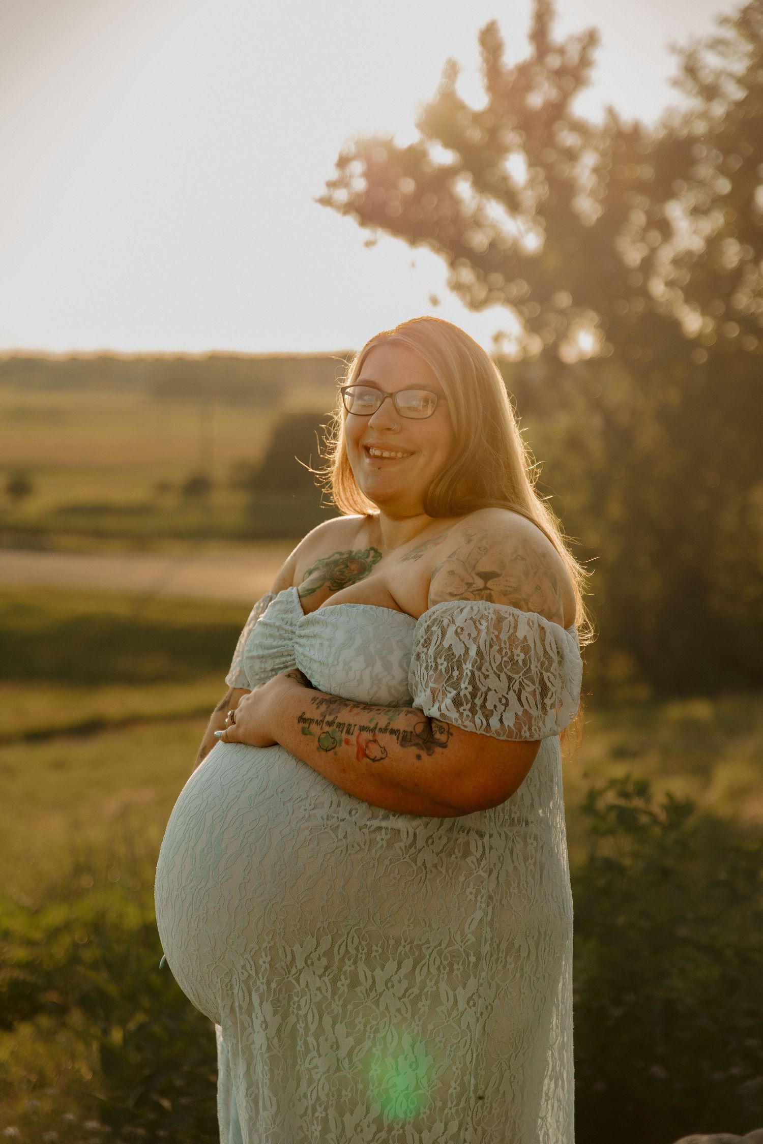 Smiling pregnant woman in white lace off-shoulder dress hugging her belly outdoors at golden hour sunset.