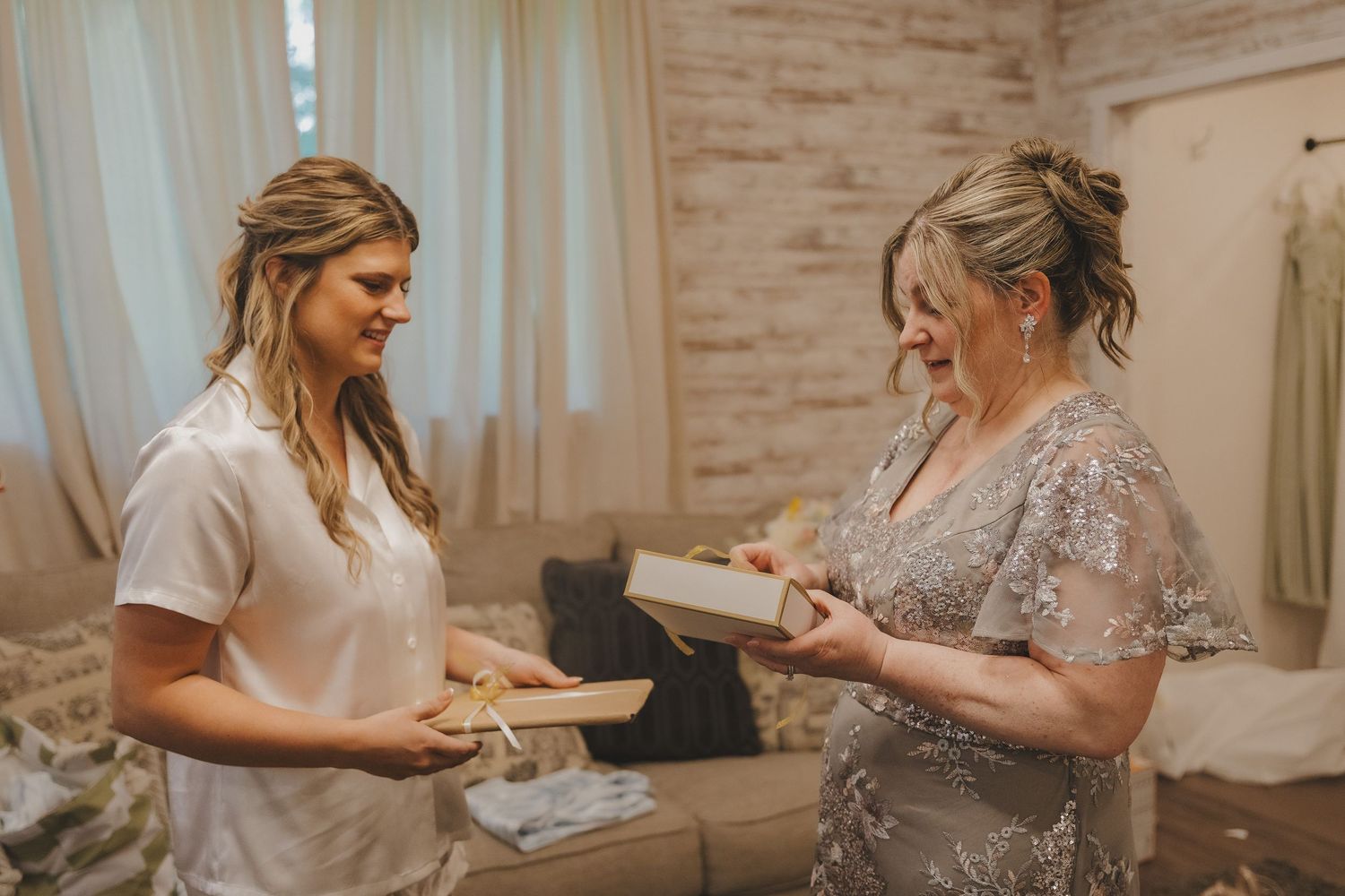 Two women exchanging gifts in a bridal suite, one in white robe and other in silver gown.