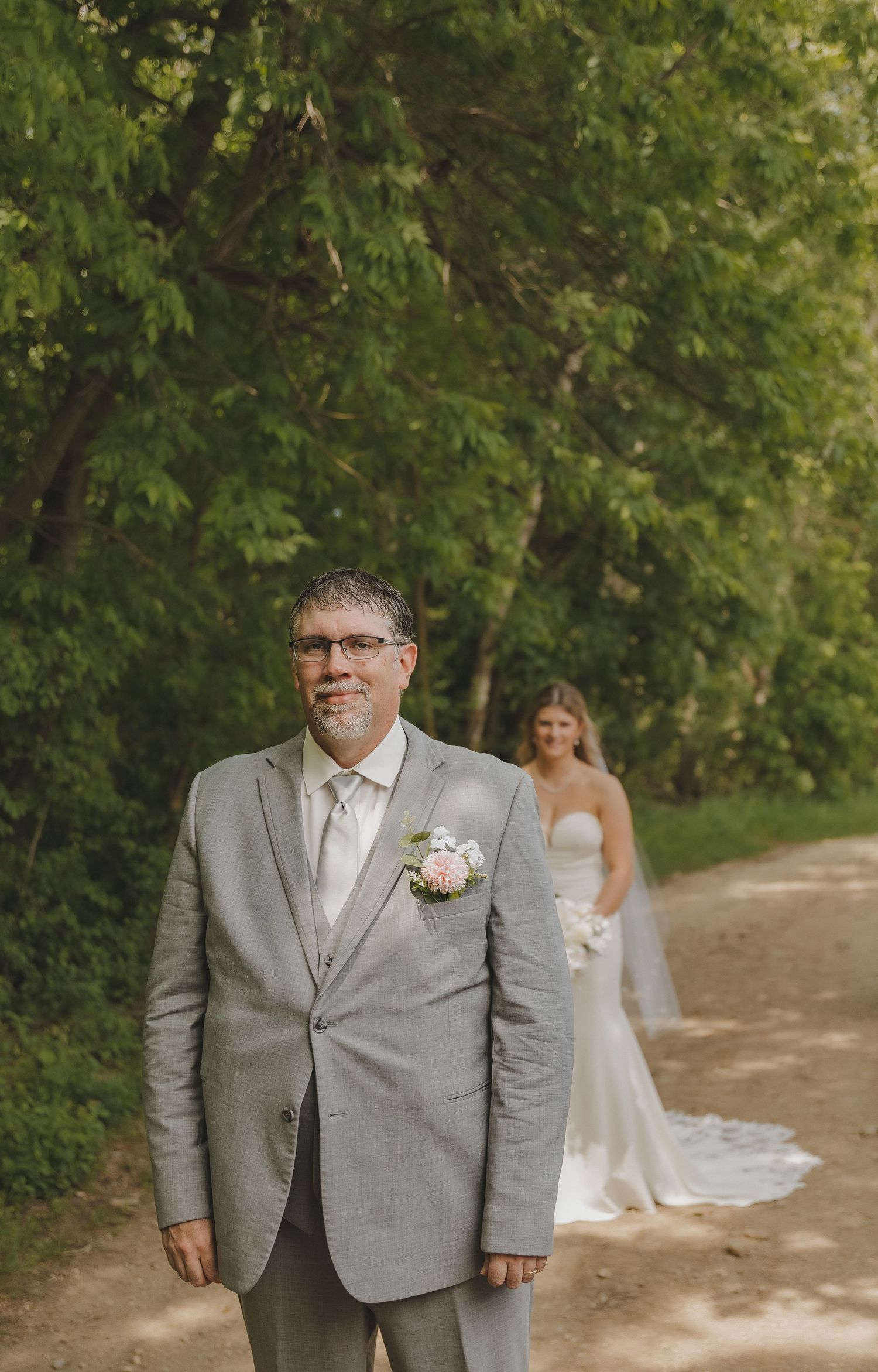 A man in a gray suit with a boutonniere stands on a tree-lined path as a bride in white approaches behind him.