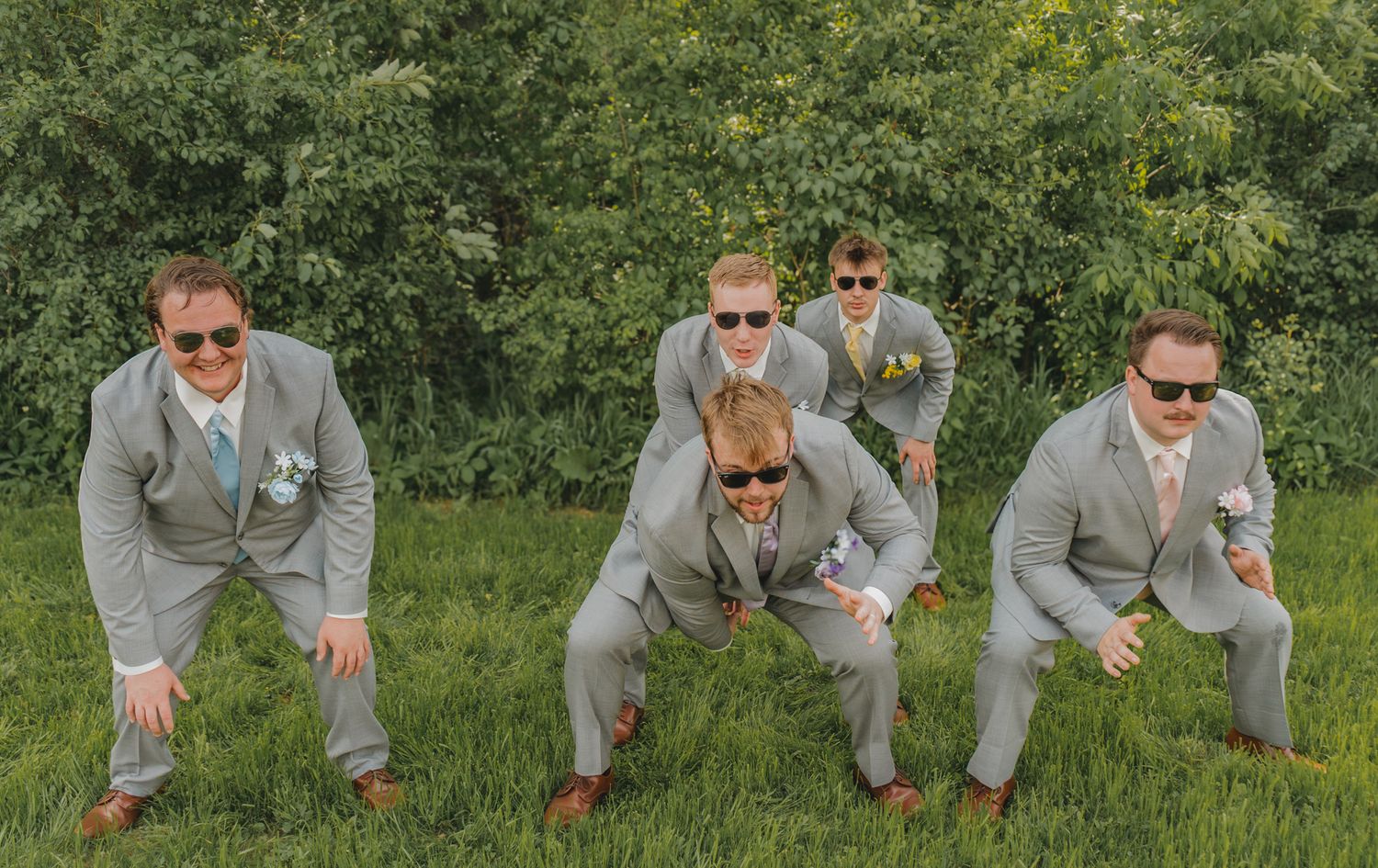 Five groomsmen in matching gray suits and sunglasses striking fun poses on a green lawn at a wedding.