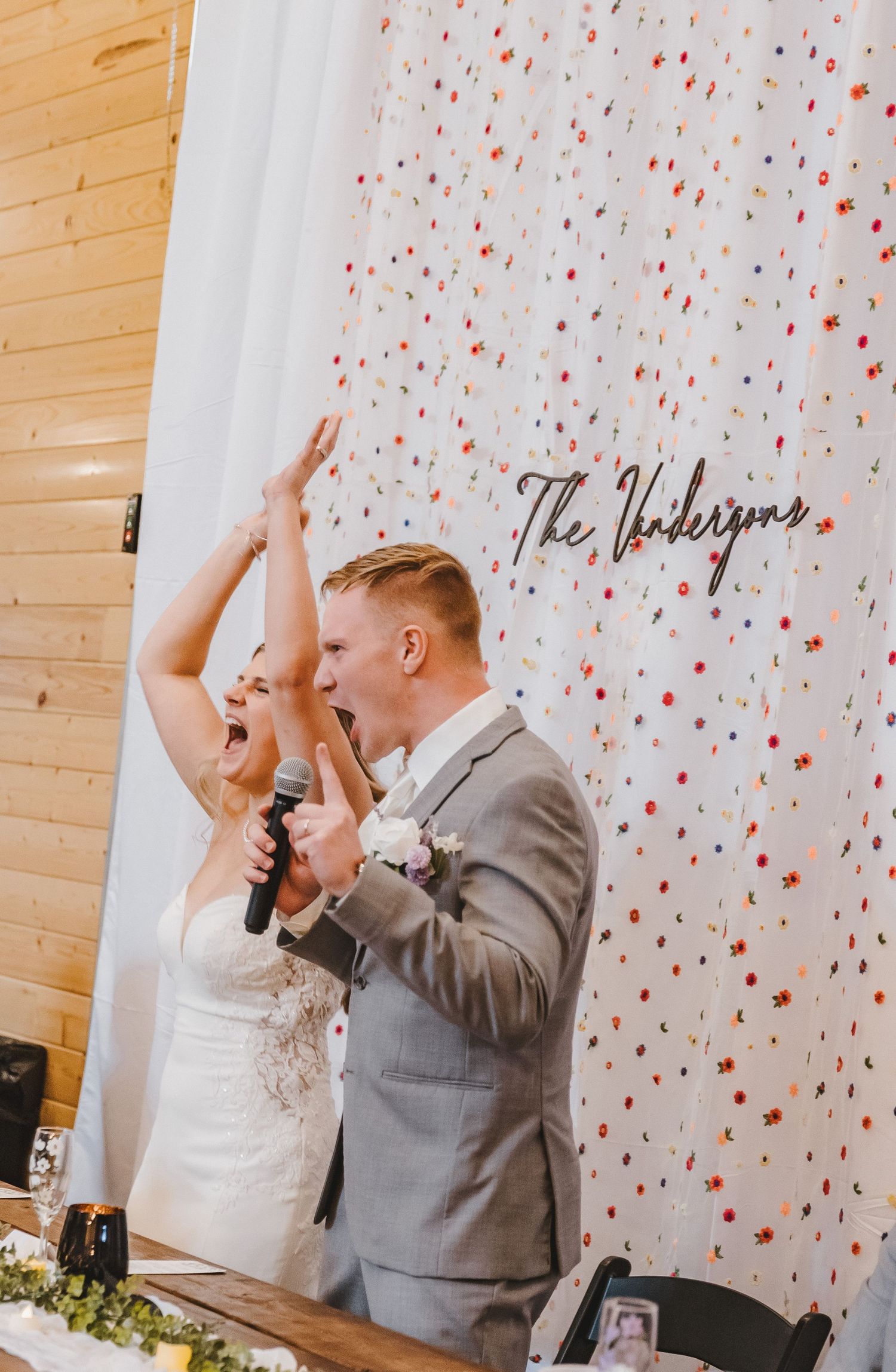 Excited bride and groom celebrate at reception table in front of floral backdrop reading The Vandersons.