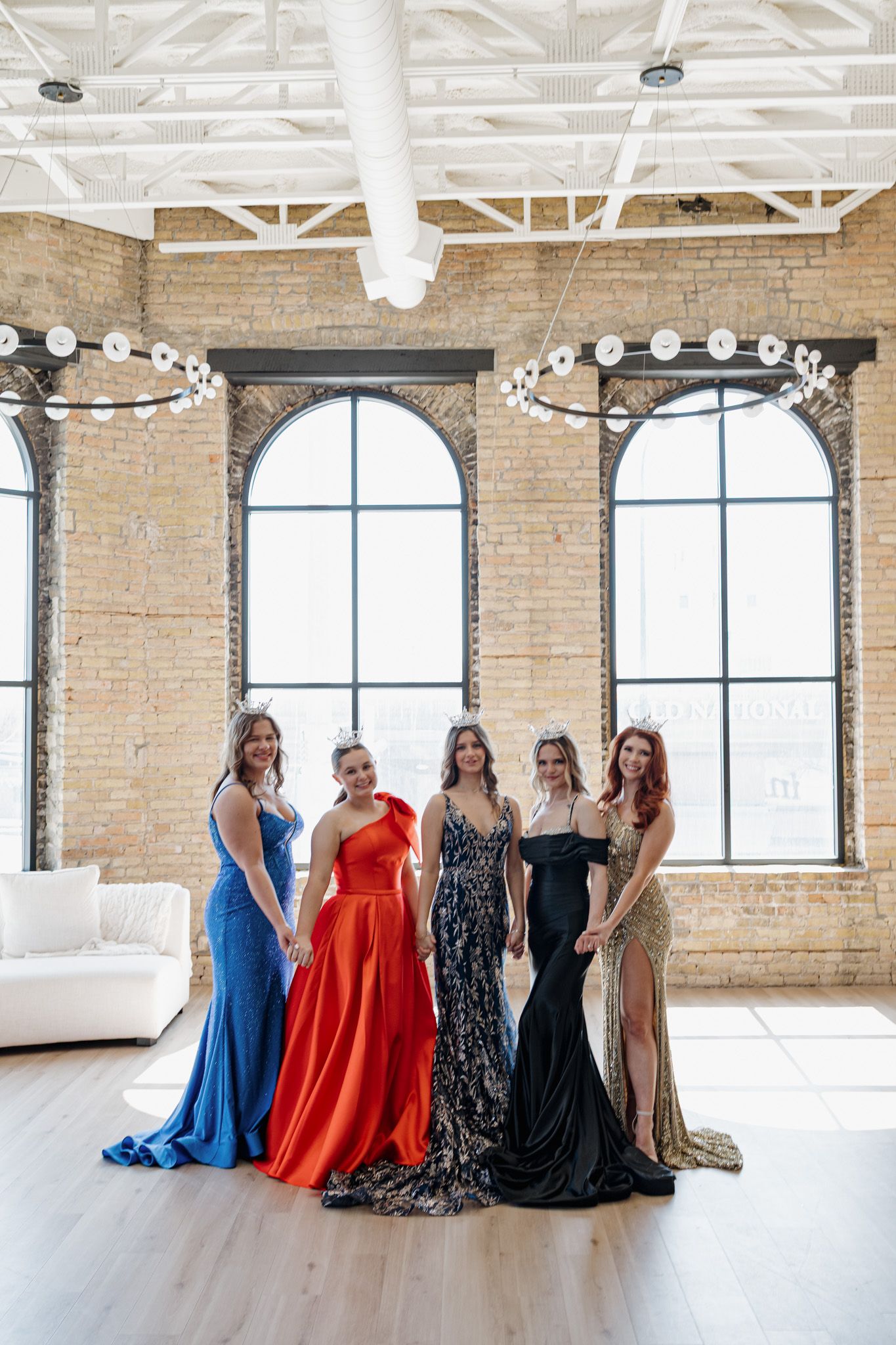 Five women in elegant formal gowns pose together in a bright loft venue with exposed brick walls and arched windows.