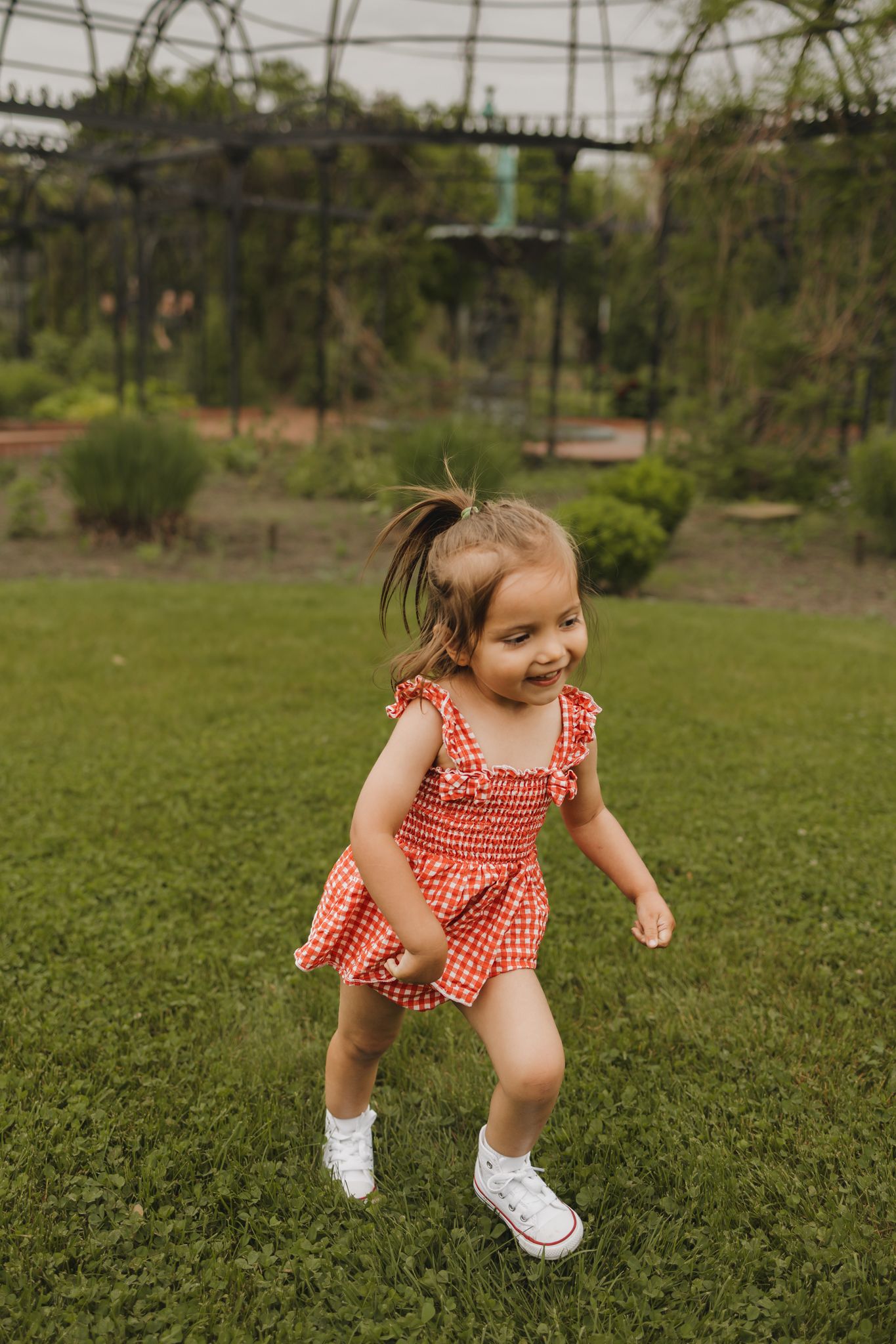 A cheerful toddler girl in a red gingham dress and white sneakers runs and plays on green grass in a garden.