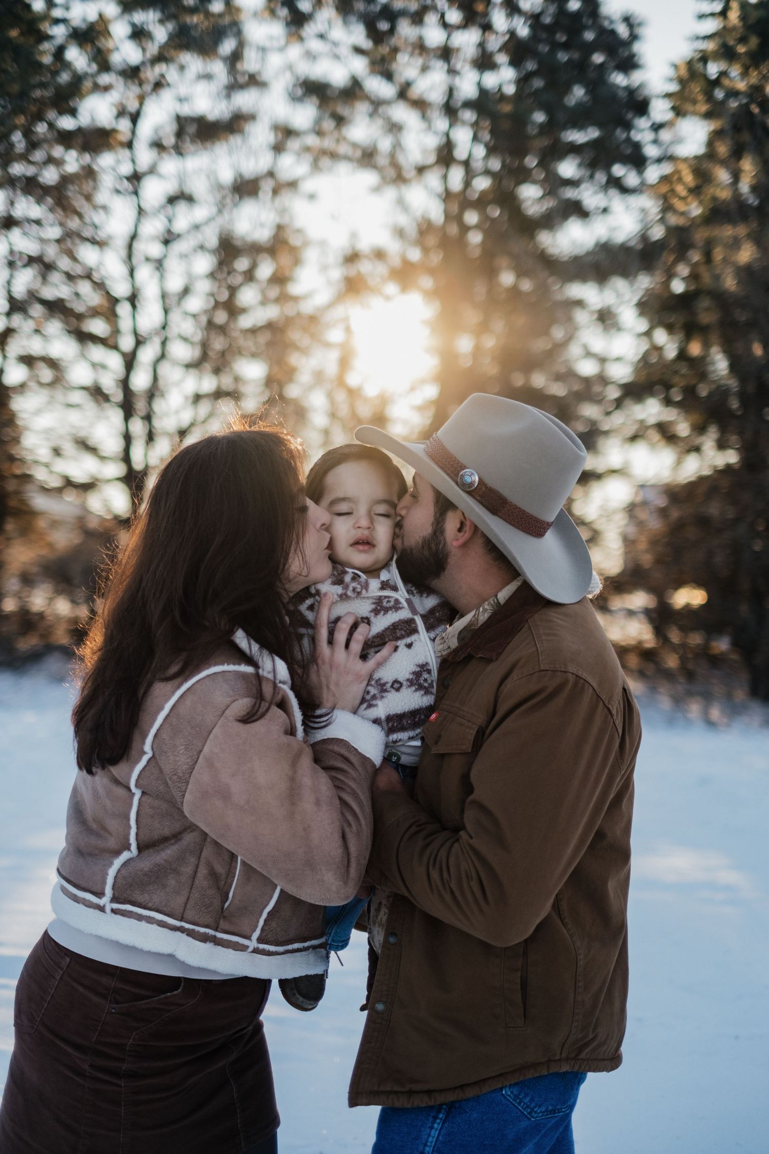 Sweet family moment captured in snowy setting with pine trees and golden sunlight.