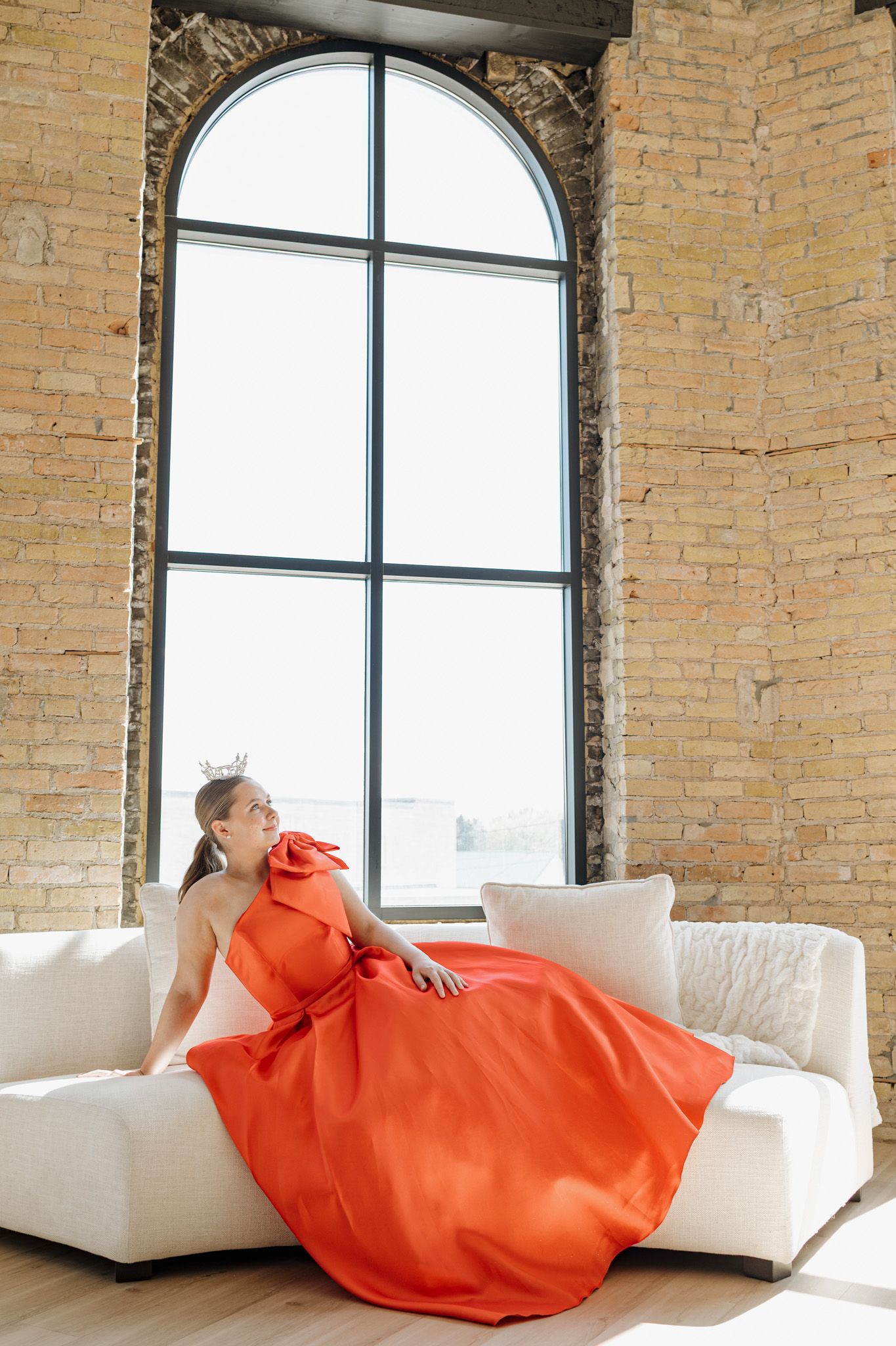 Woman in elegant orange gown seated on white sofa before arched window in brick loft.