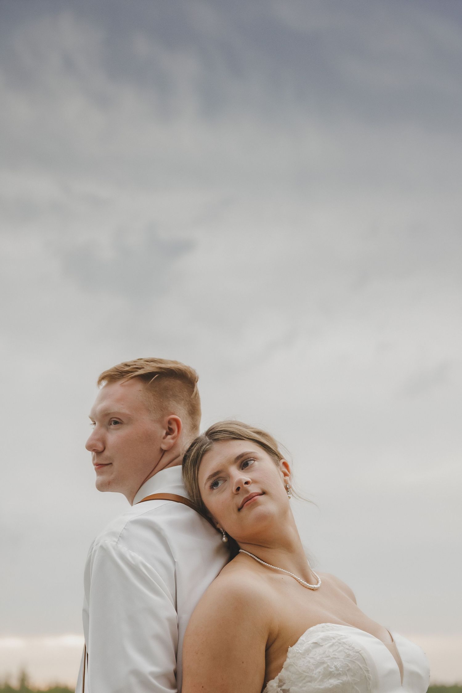 Bride and groom standing back to back against a dramatic cloudy sky in romantic wedding portrait.