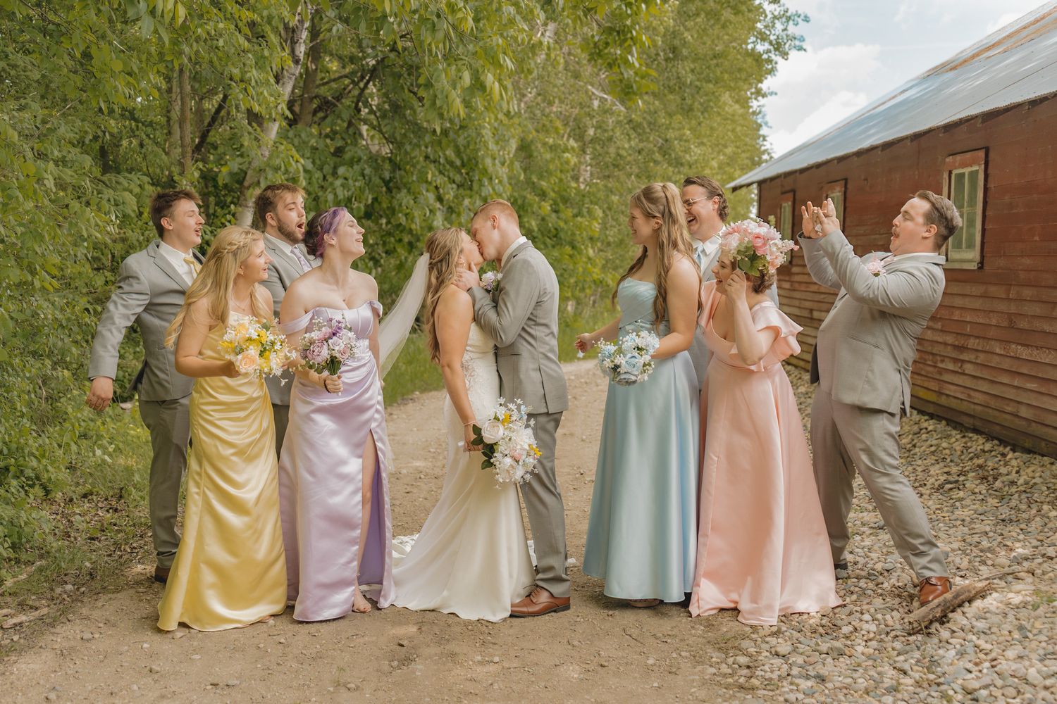 Wedding party poses outdoors, bride and groom kissing, bridesmaids in colorful pastel gowns, groomsmen in gray suits.