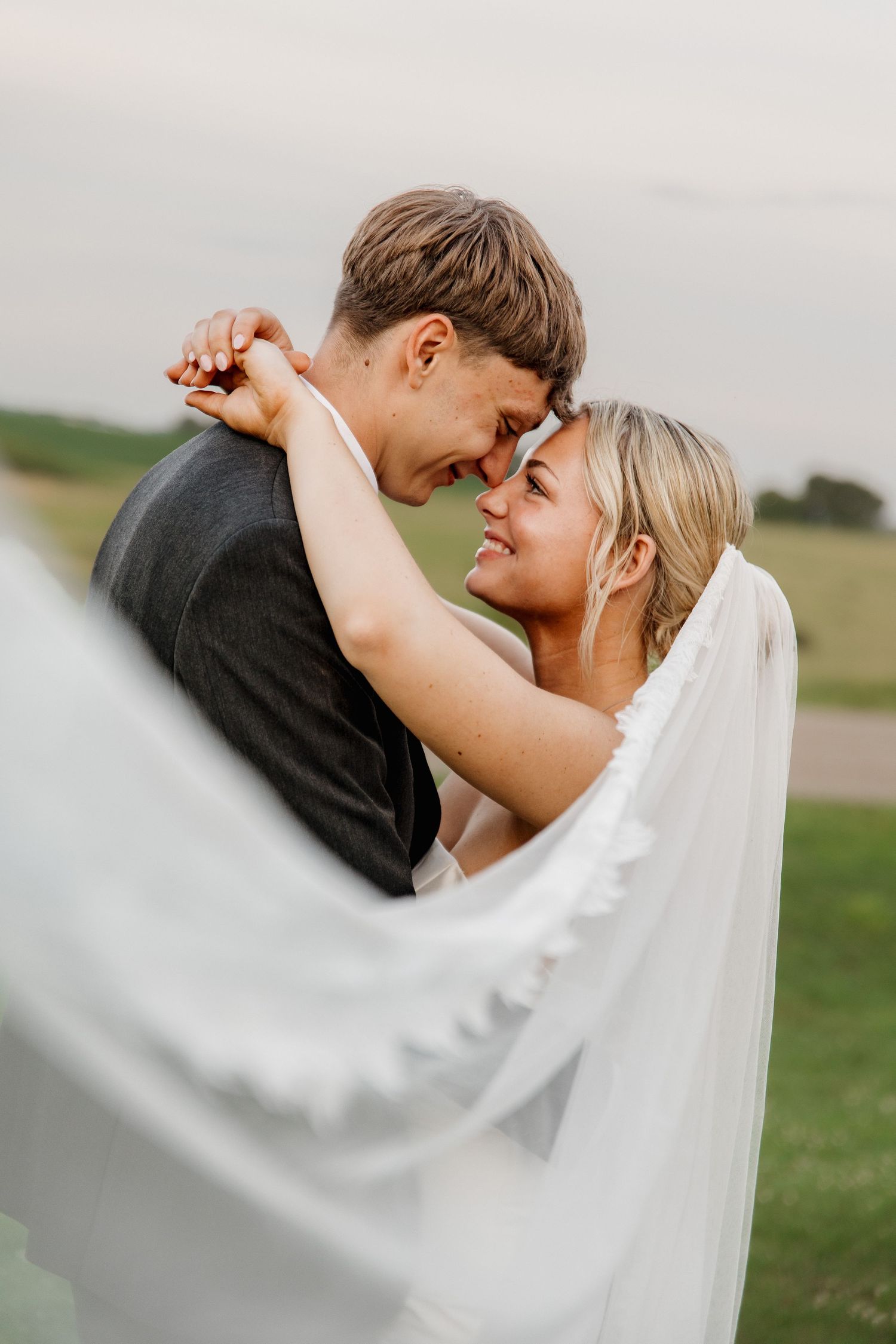 Happy bride smiles at groom touching foreheads together outdoors, white veil flowing in foreground at golden hour.