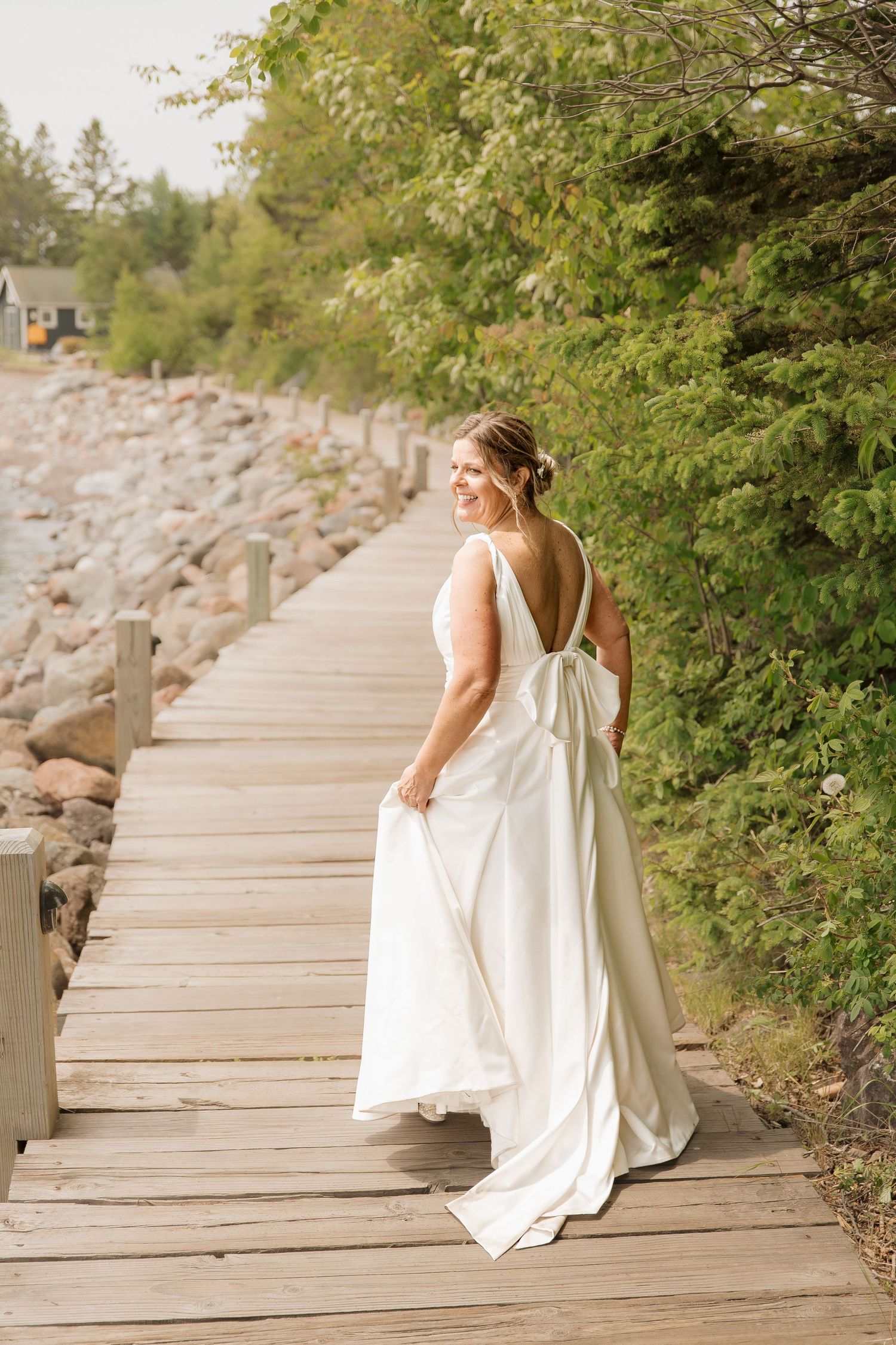 Bride in white gown walks on wooden boardwalk by rocky shore, glancing back with a smile.