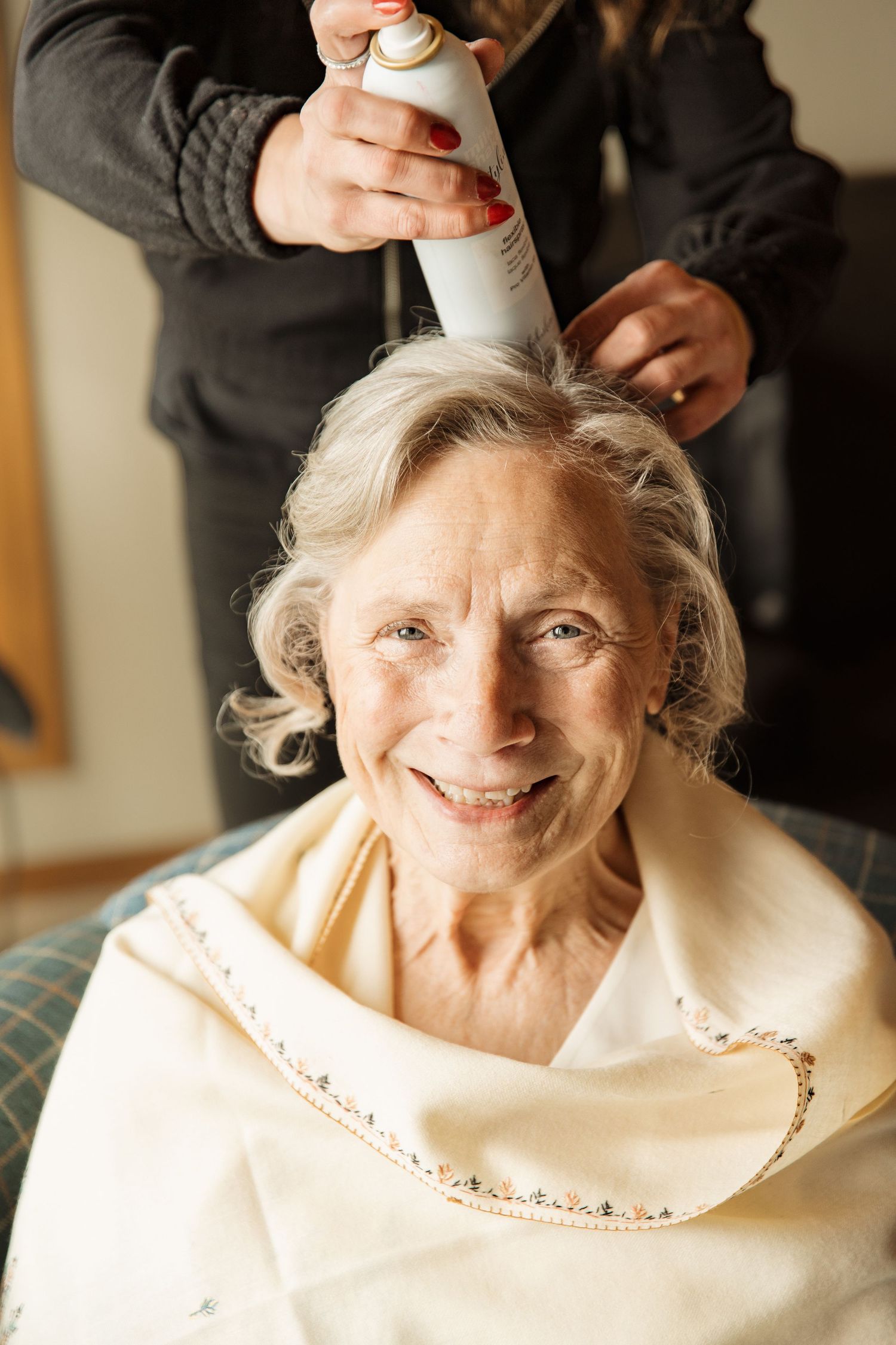 Smiling elderly woman getting hair styled with spray at a salon appointment.