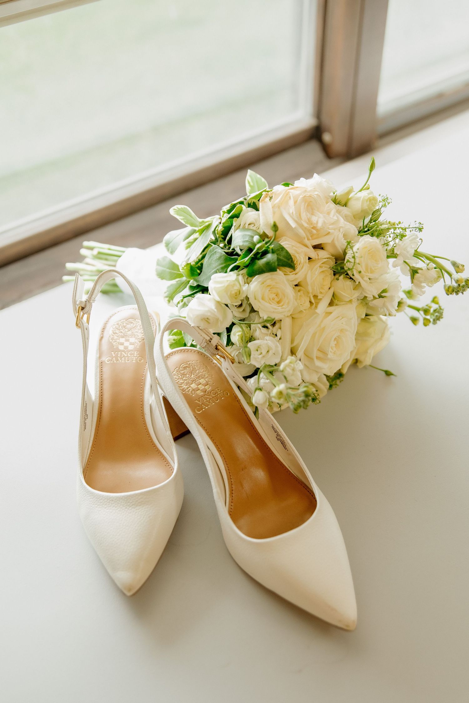 White satin bridal heels and elegant white rose bouquet displayed on windowsill in natural light.