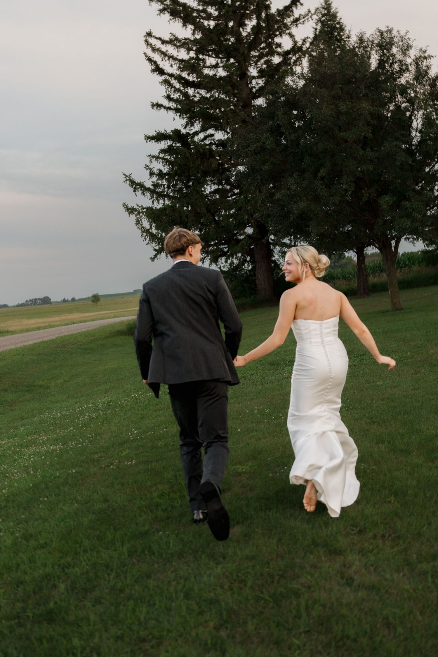 Bride and groom holding hands running across a green lawn at dusk during their outdoor wedding celebration.