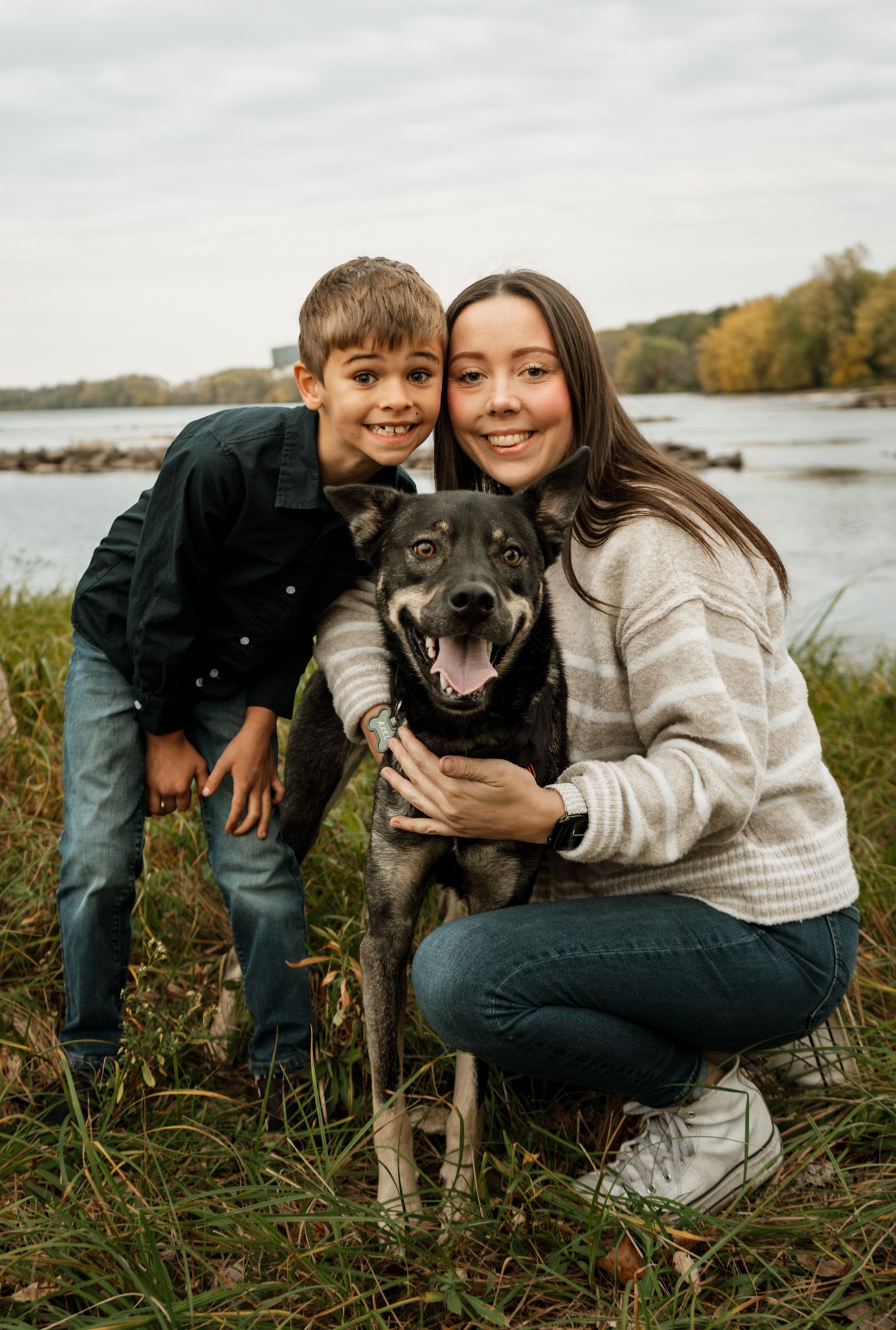 Family and their black dog posing together by a lakeside on an autumn day.