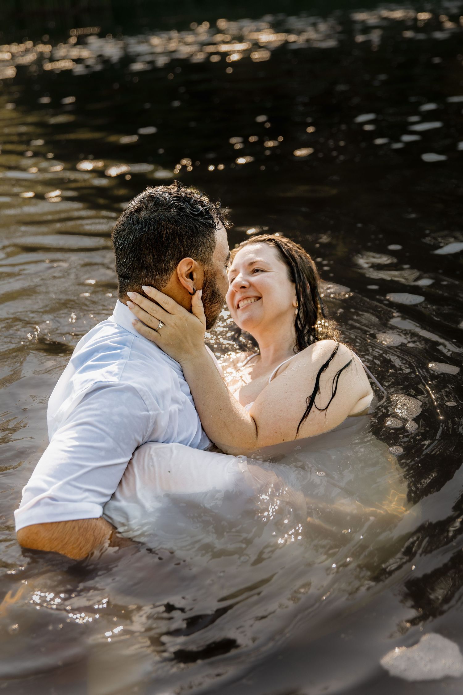 A couple in wedding attire sharing a romantic moment while wading in a sunlit lake.