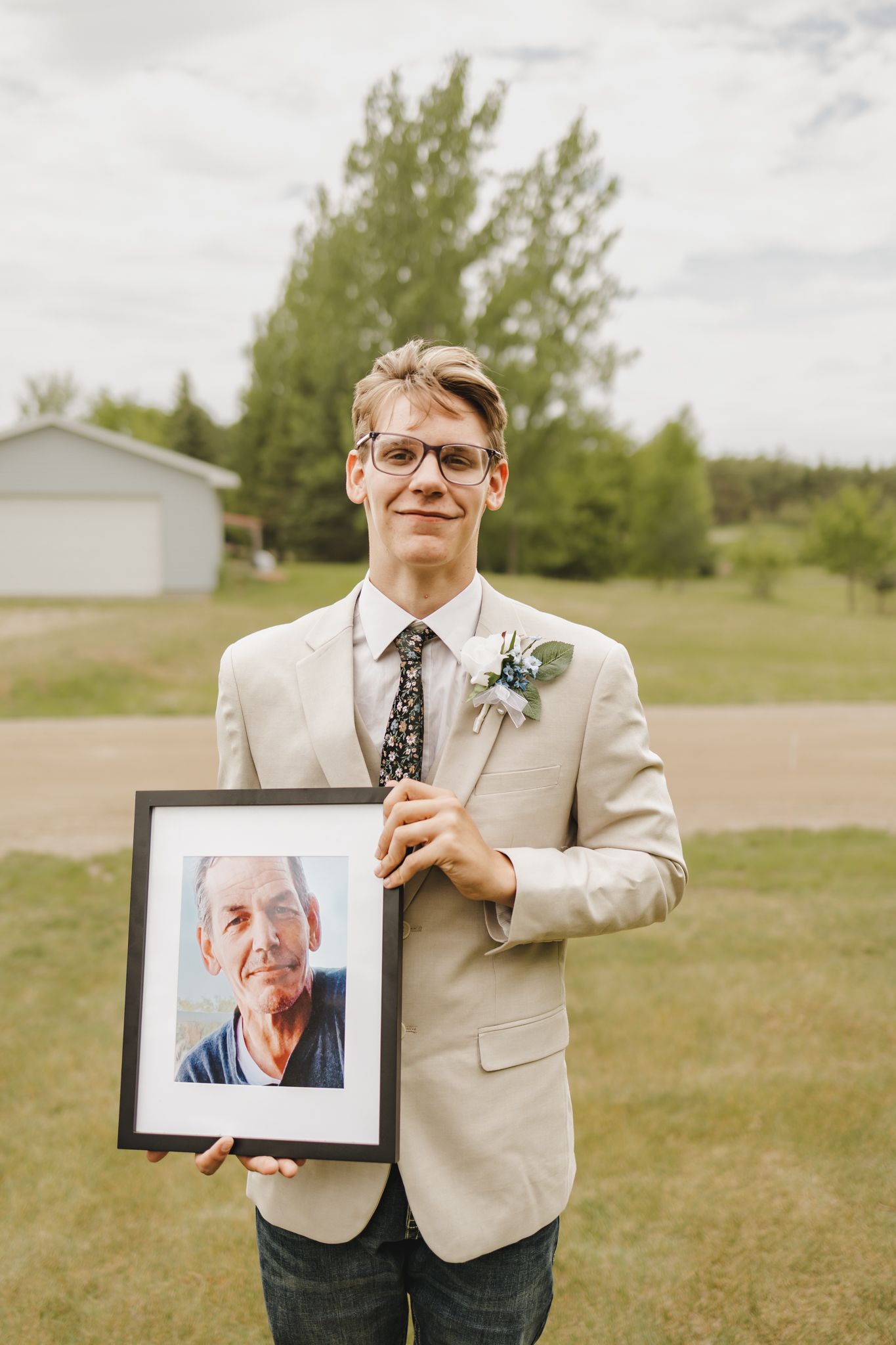 Groom in beige suit holding a framed memorial photo with a white boutonniere at a wedding.