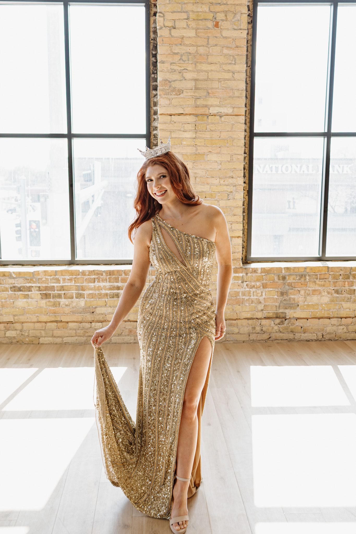 Woman in gold sequin one-shoulder gown with slit poses in sunlit brick loft space.