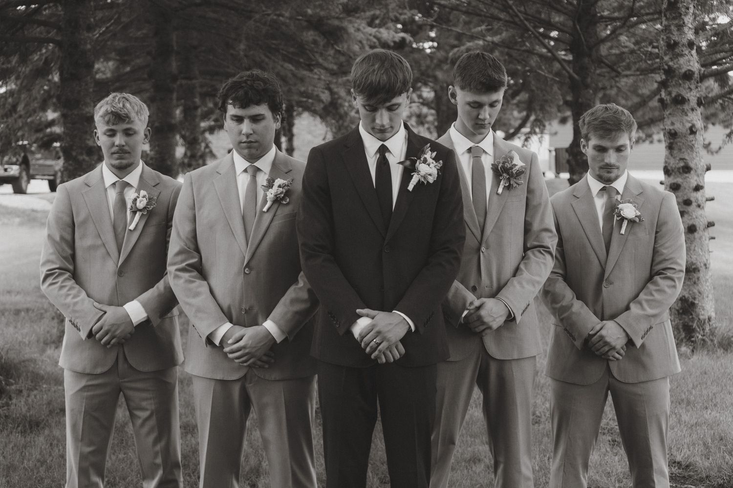 Black and white photo of a groom and four groomsmen in suits with boutonnieres standing outdoors at a wedding.