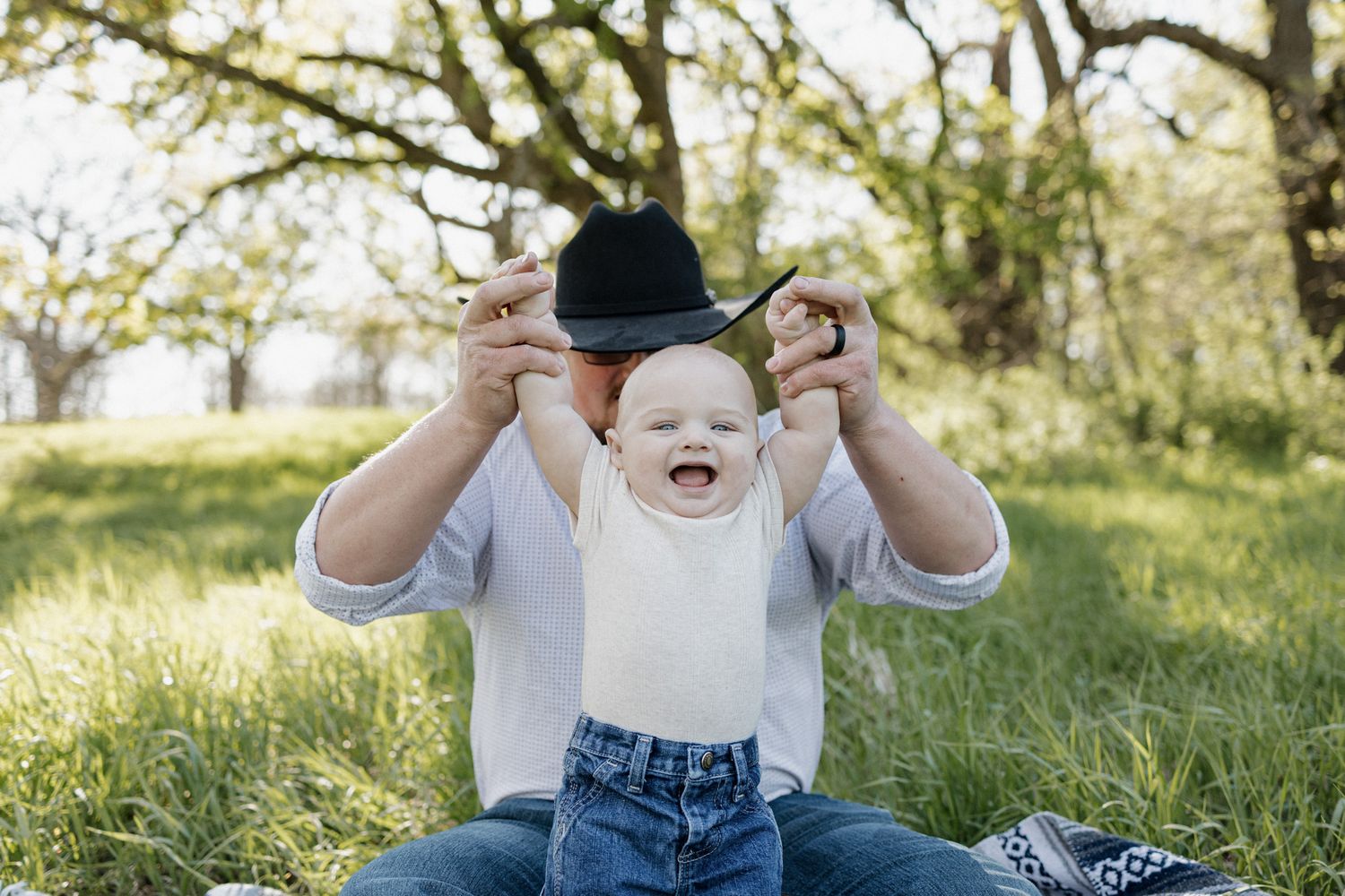 A laughing baby wearing a bib sits on grass as an adult playfully holds a black cowboy hat above their head outdoors.