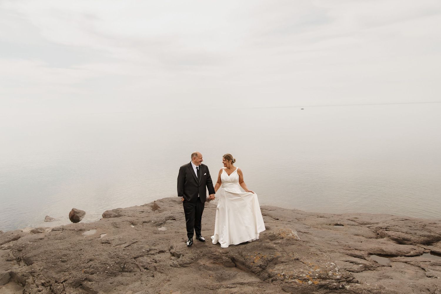 Bride and groom hold hands walking on rocky shoreline with misty lake backdrop on their wedding day.