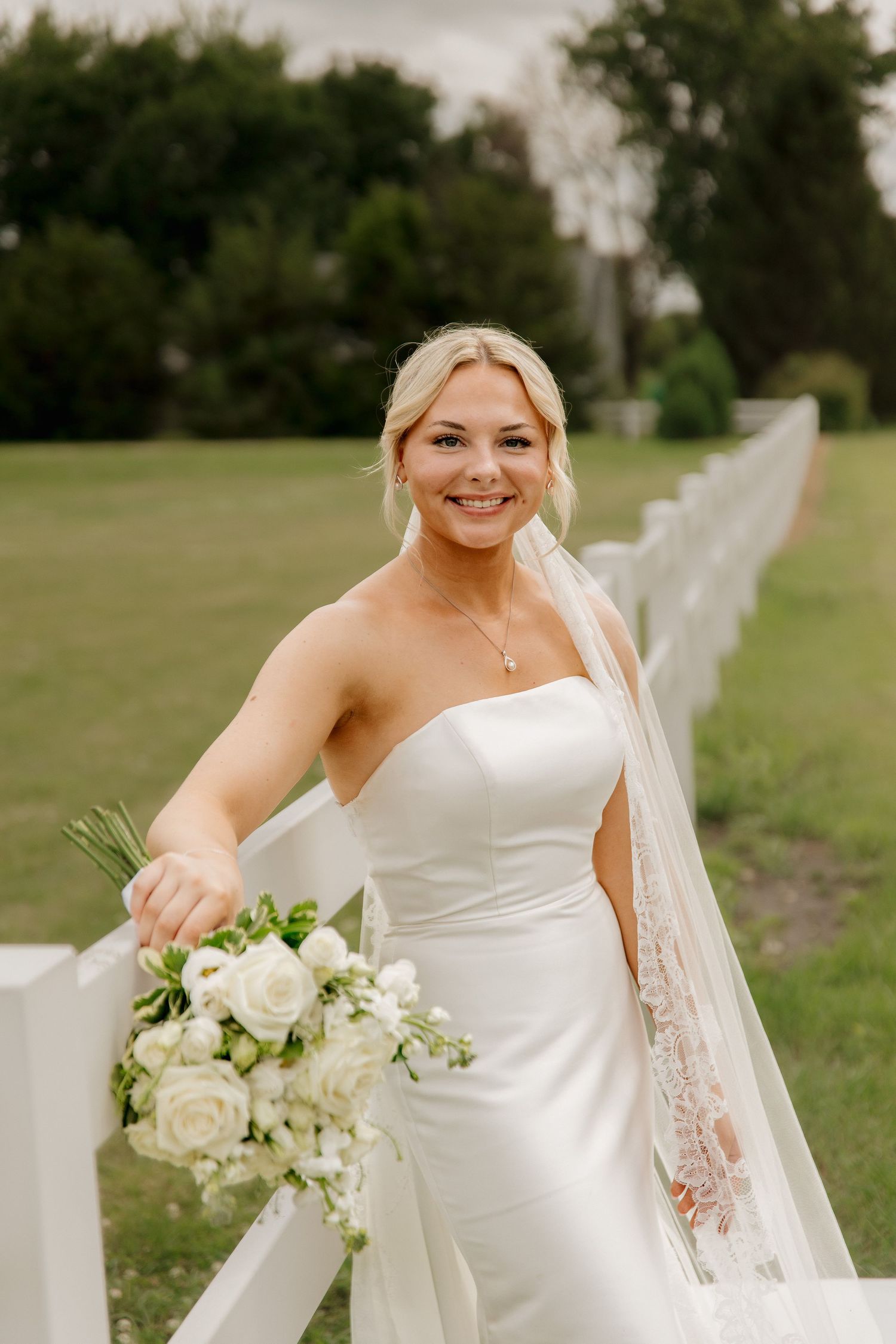 Smiling blonde bride in strapless gown with lace veil holding white rose bouquet by white fence.