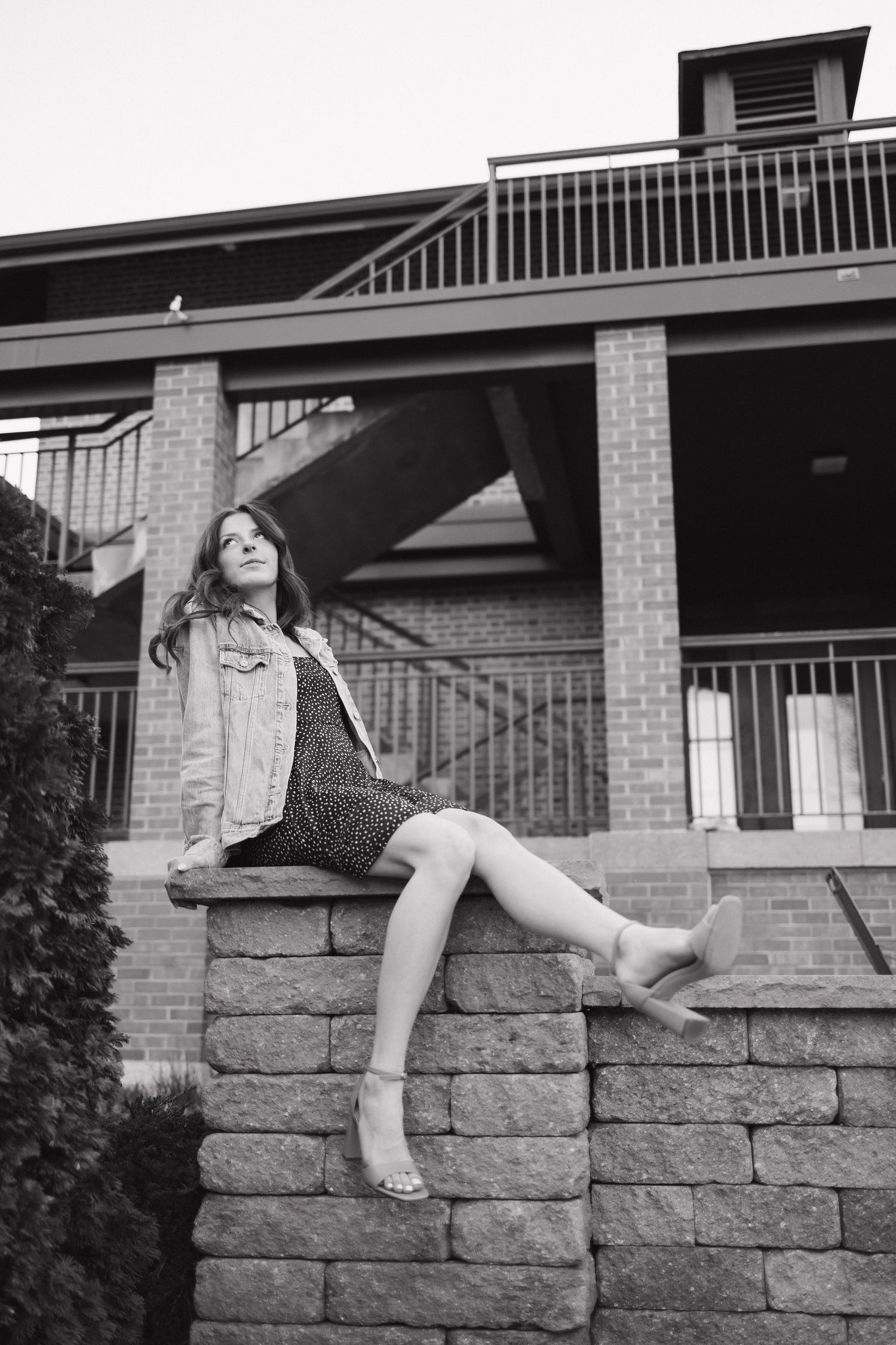 Black and white photo of a young woman in a dress and jacket sitting on a stone wall outside a brick building.