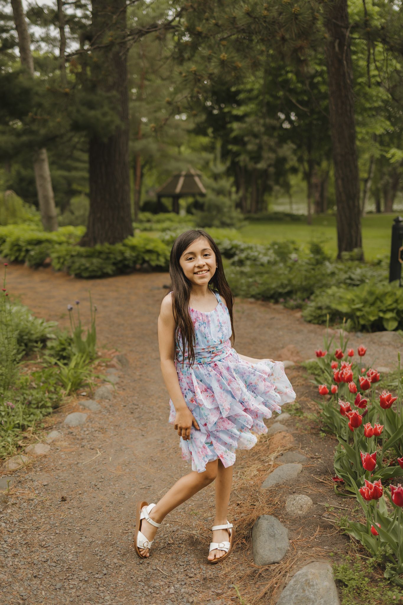 A smiling young girl in a floral dress twirls on a garden path surrounded by red tulips and lush green trees.