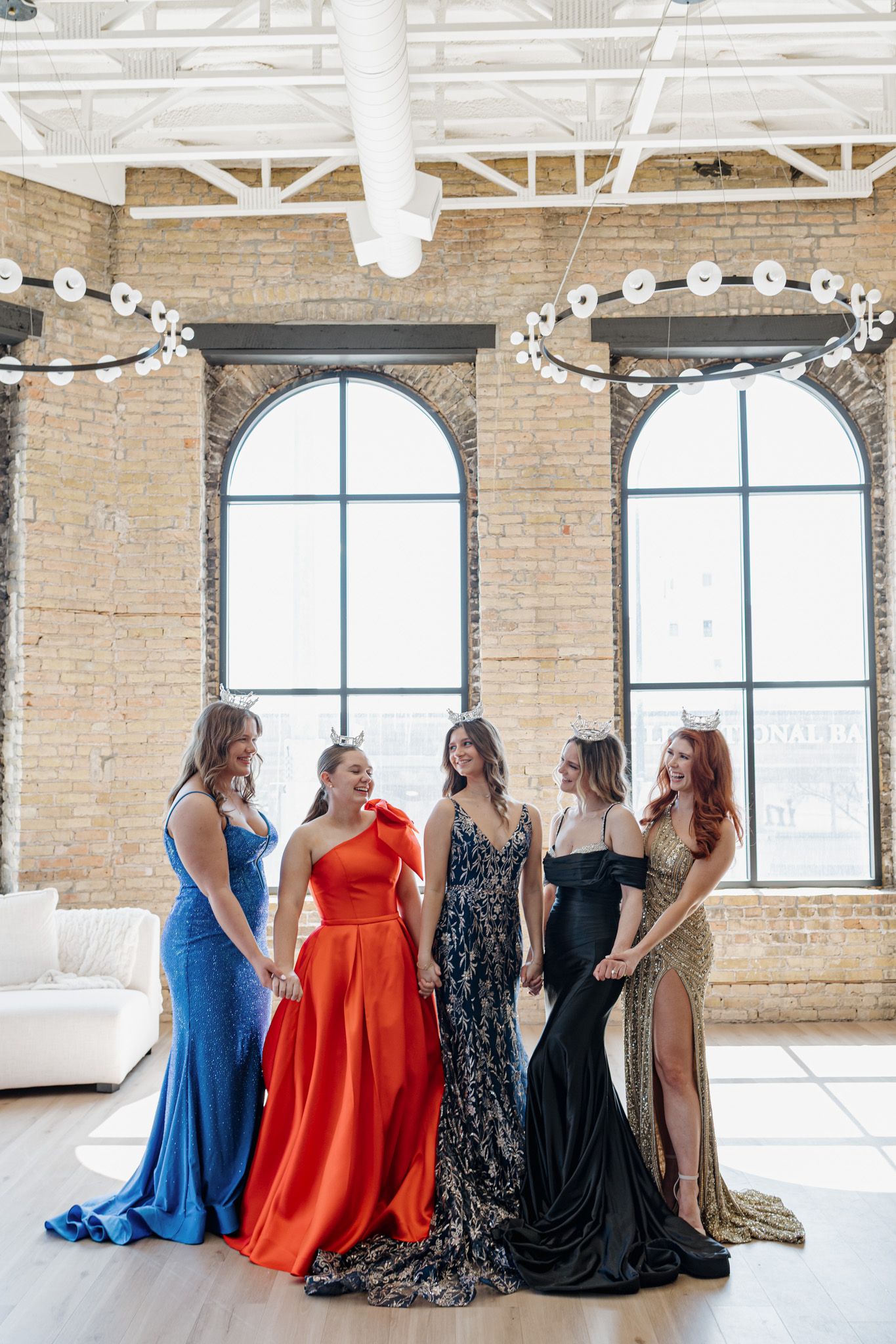 Five women in formal gowns laughing together in industrial brick loft with arched windows.