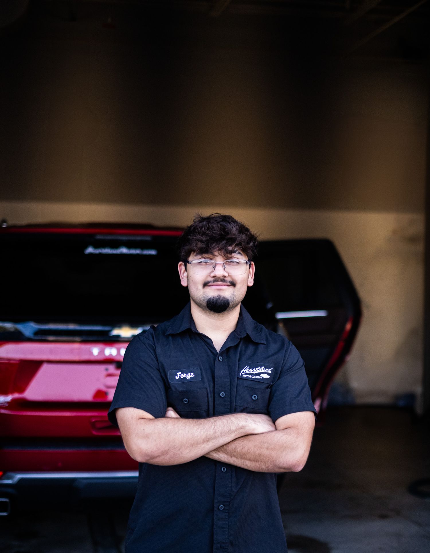 Auto technician in black uniform standing with crossed arms in front of red vehicle.