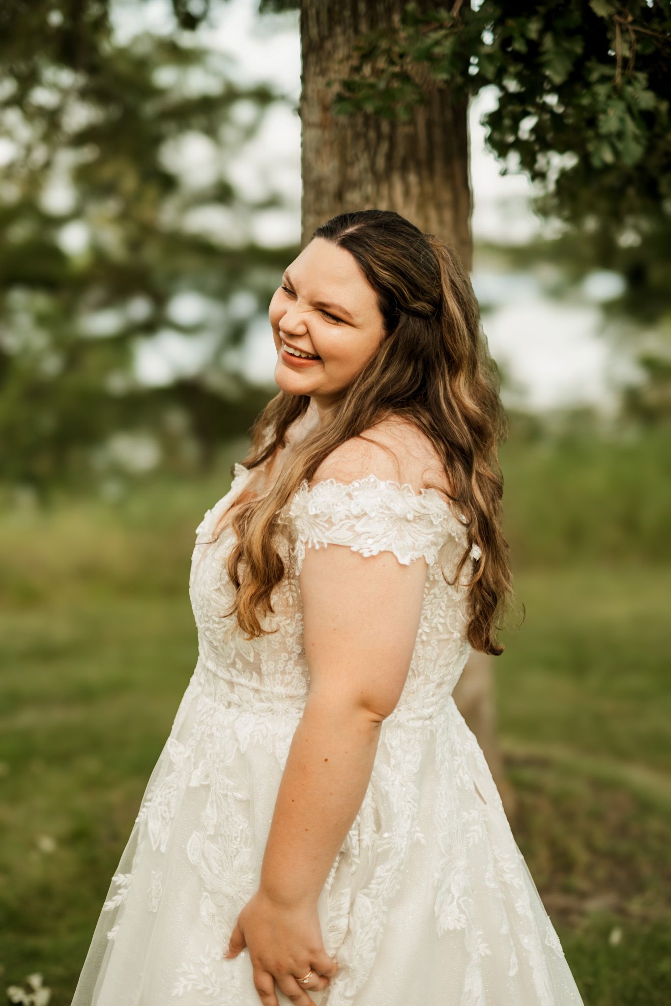 A bride in an off-shoulder lace wedding dress smiles joyfully in an outdoor garden setting.