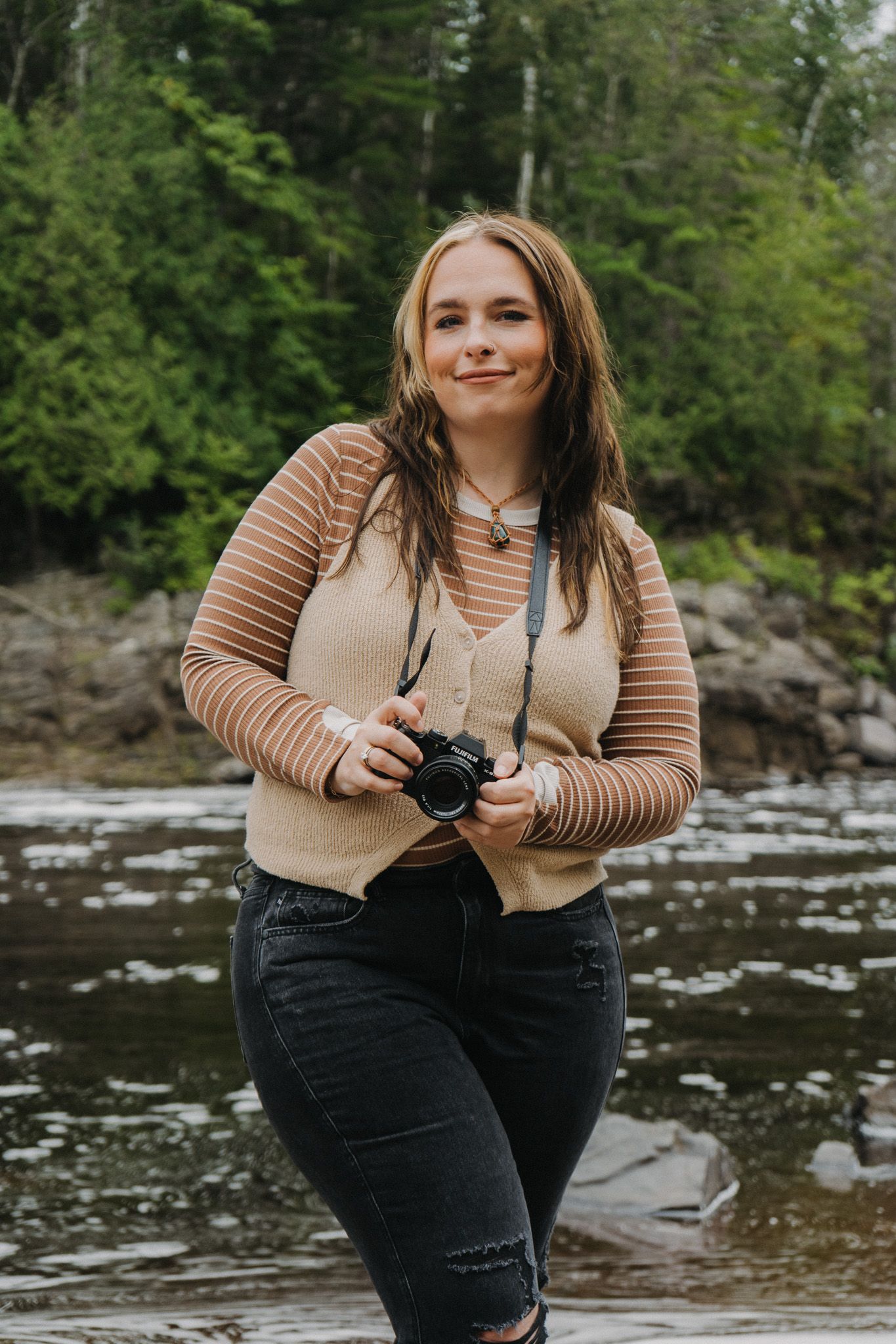 Woman with camera sits by rocky riverbank in casual autumn attire.