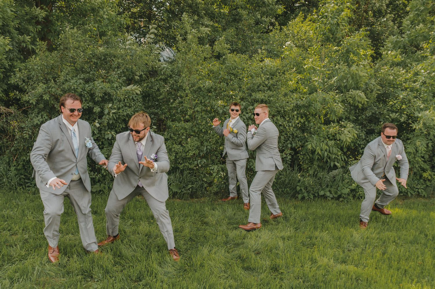Five groomsmen in gray suits dancing and celebrating outdoors in front of lush green bushes.