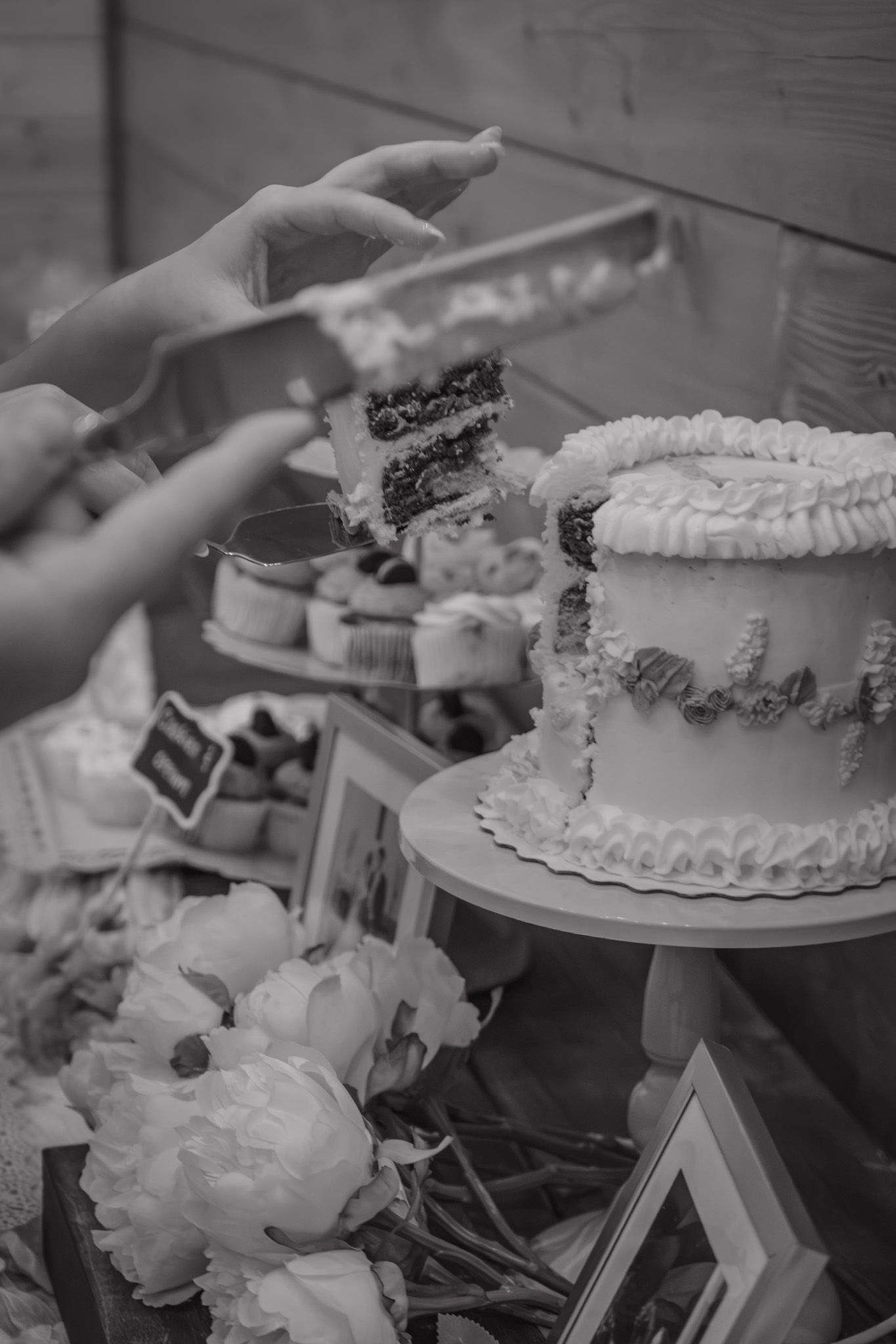 Black and white photo of wedding cake with floral decorations and hands serving a slice.