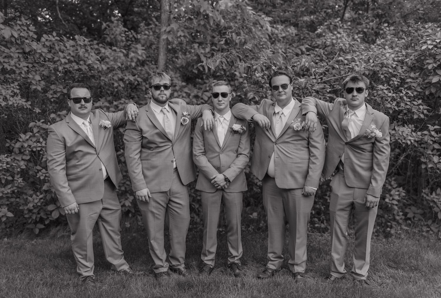 Five groomsmen in matching suits and sunglasses pose together outdoors at a wedding.